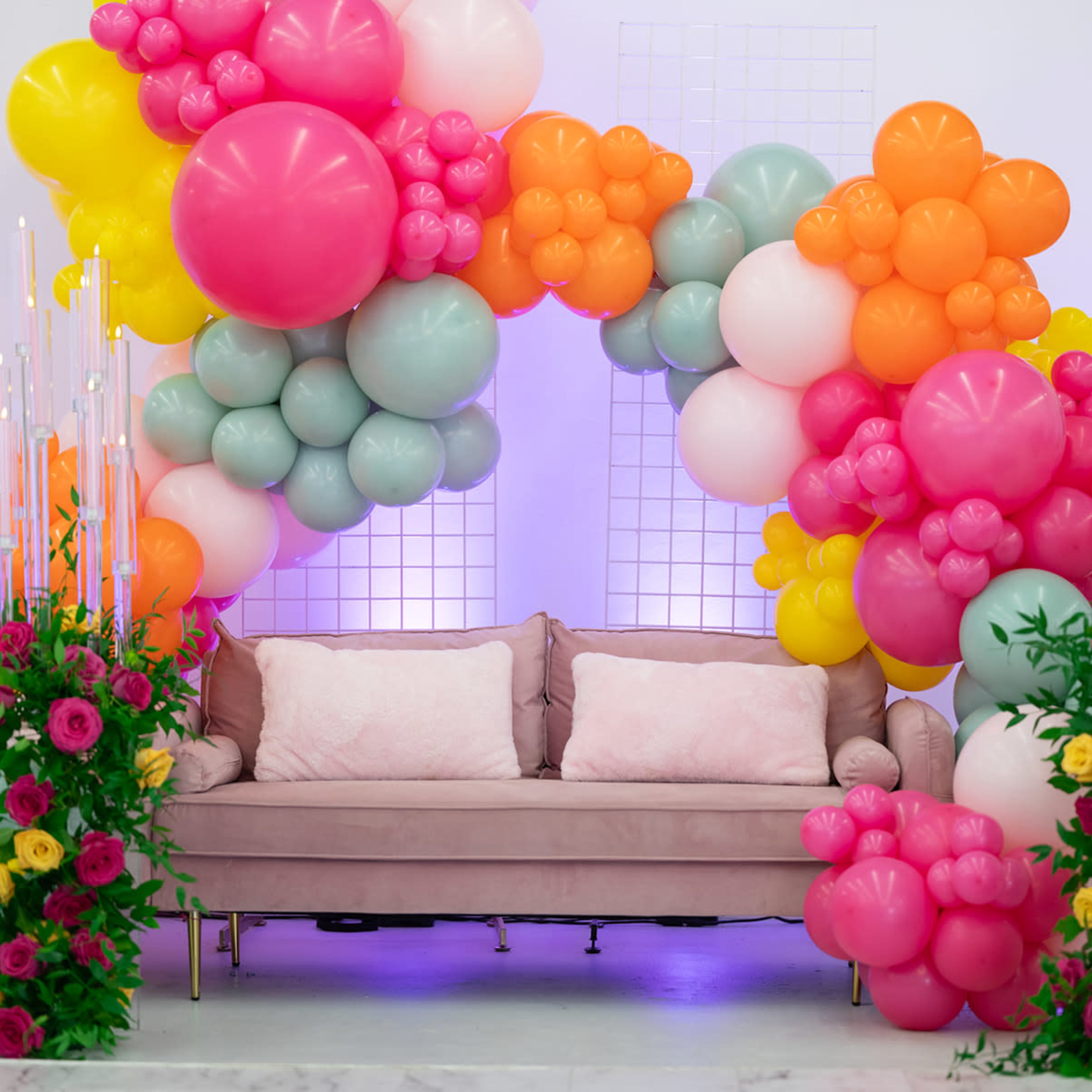 A sofa is positioned under a large arch of colorful balloons, surrounded by floral arrangements.