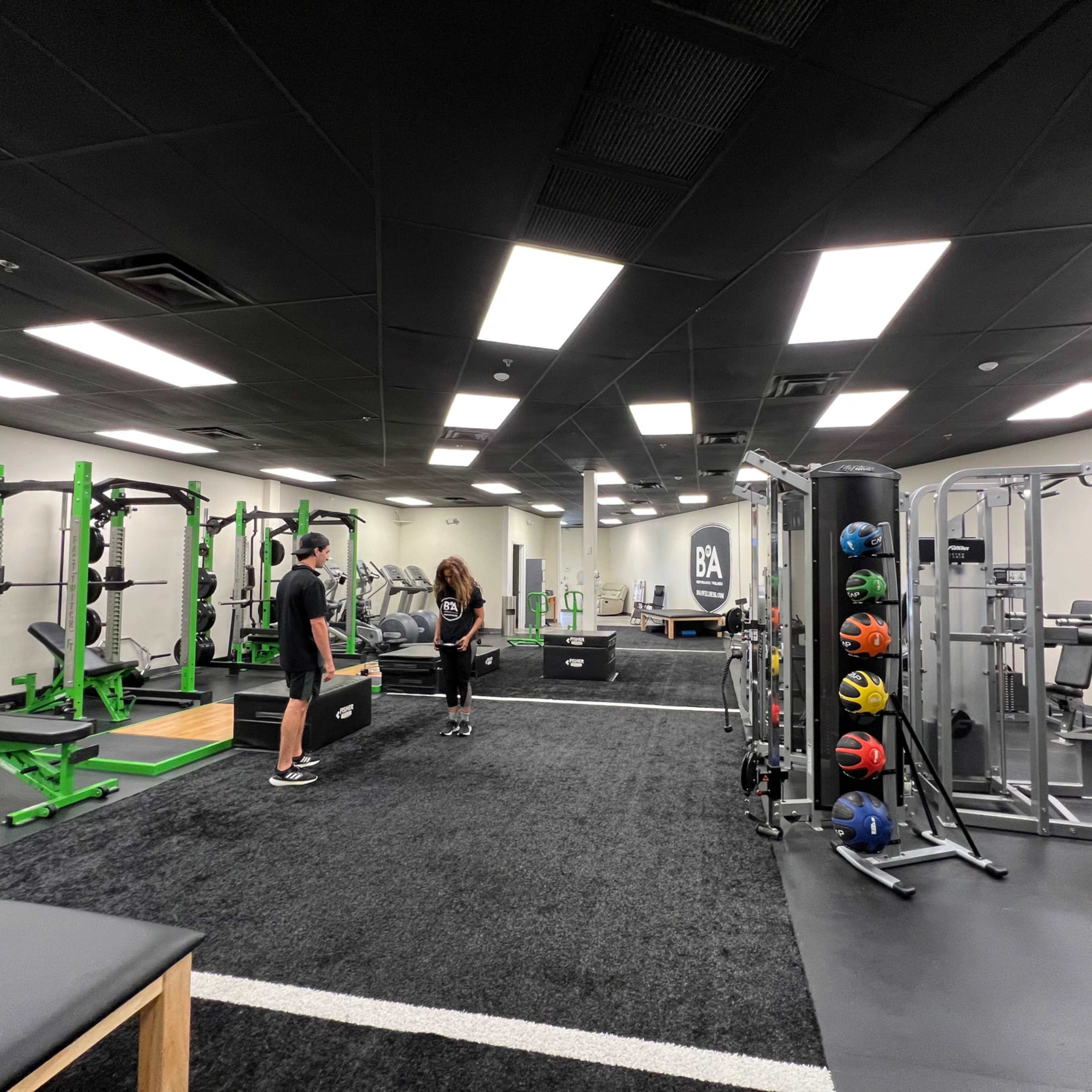 A modern gym features various exercise equipment, including weights and cardio machines, with two individuals engaged in conversation.