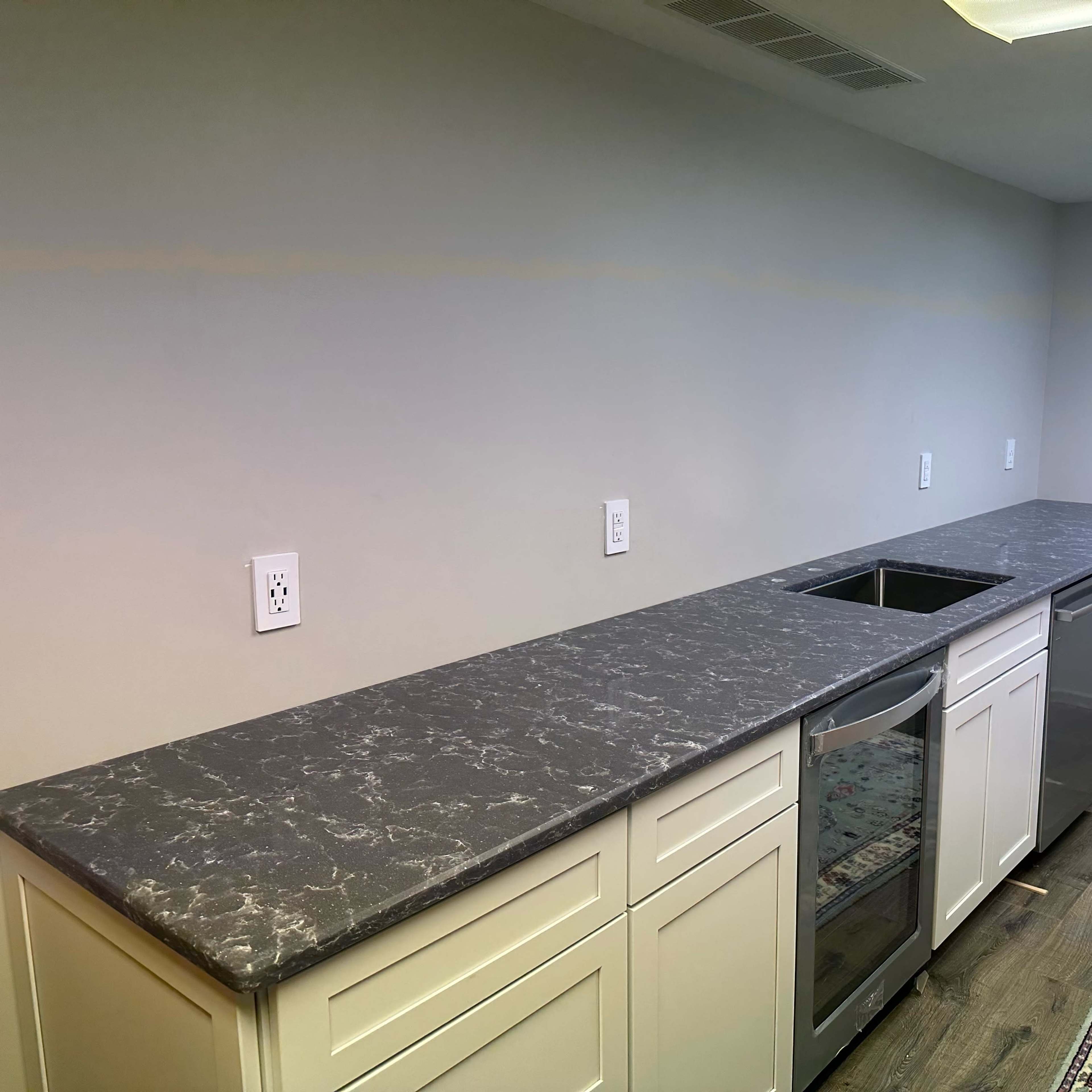 A kitchen with a long countertop, white cabinets, and a gray surface, alongside a rug on the floor.