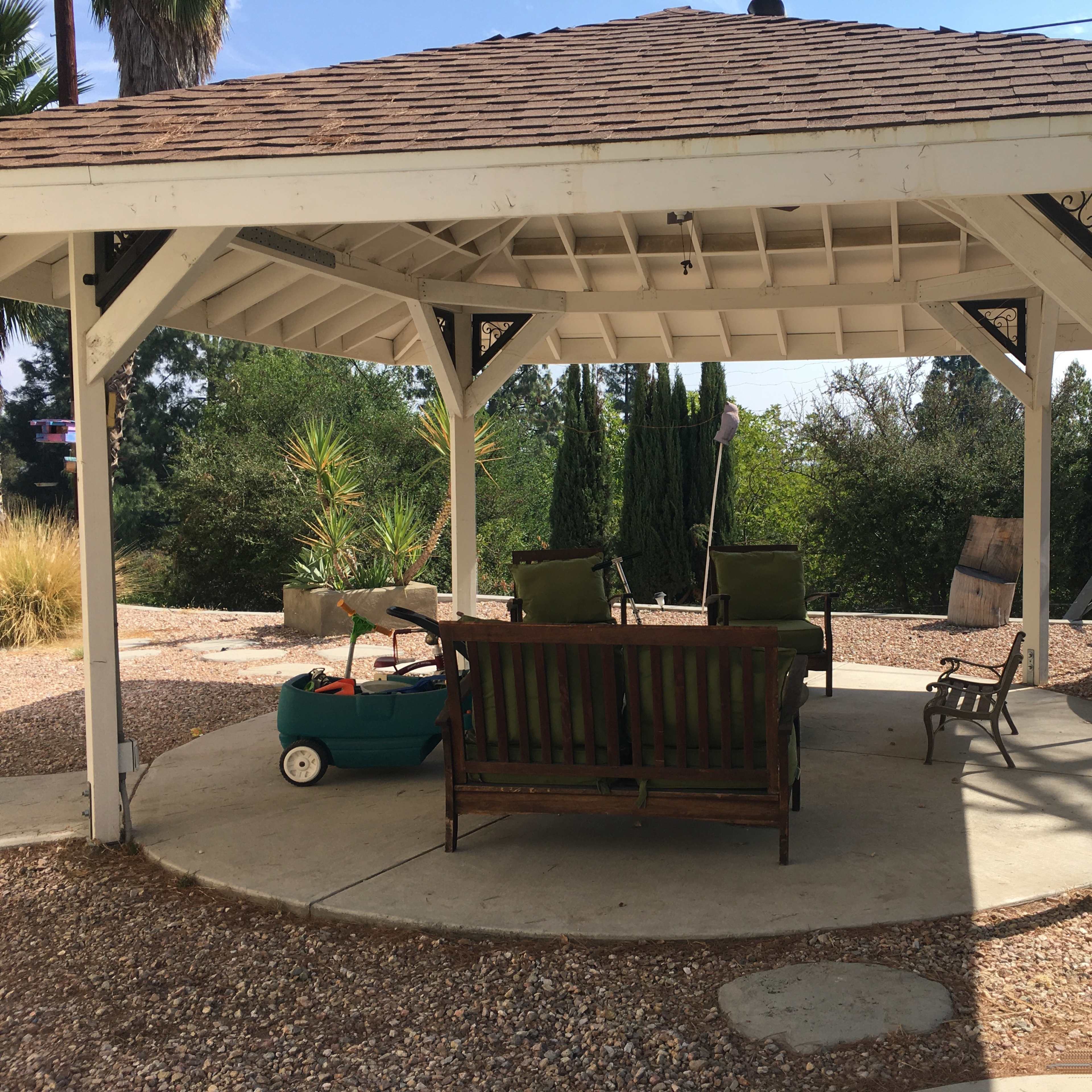A gazebo with a wooden bench and two green chairs overlooks a landscaped area with a trampoline in the background.