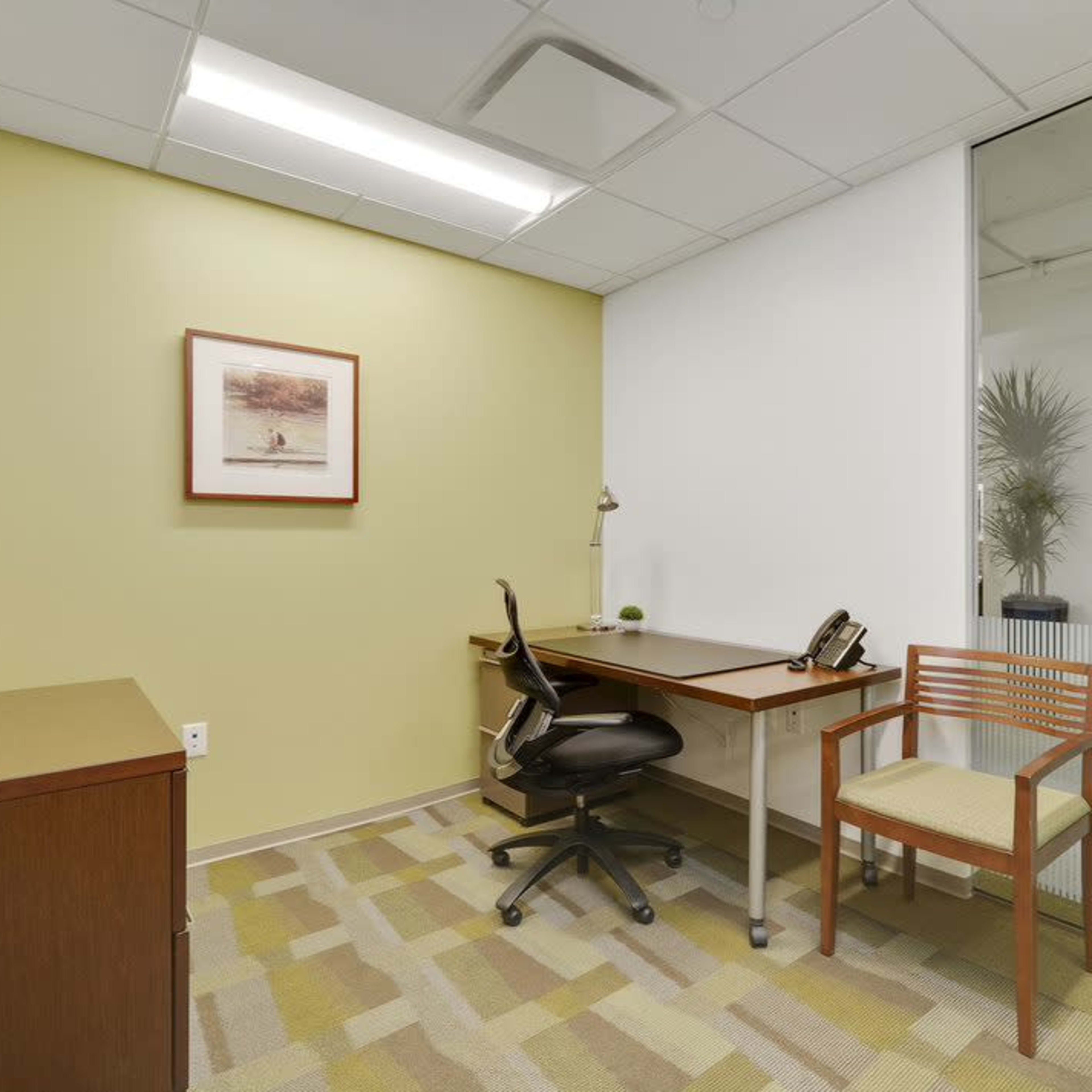 A small office space with a desk, chair, telephone, and a window featuring a view of a corridor.