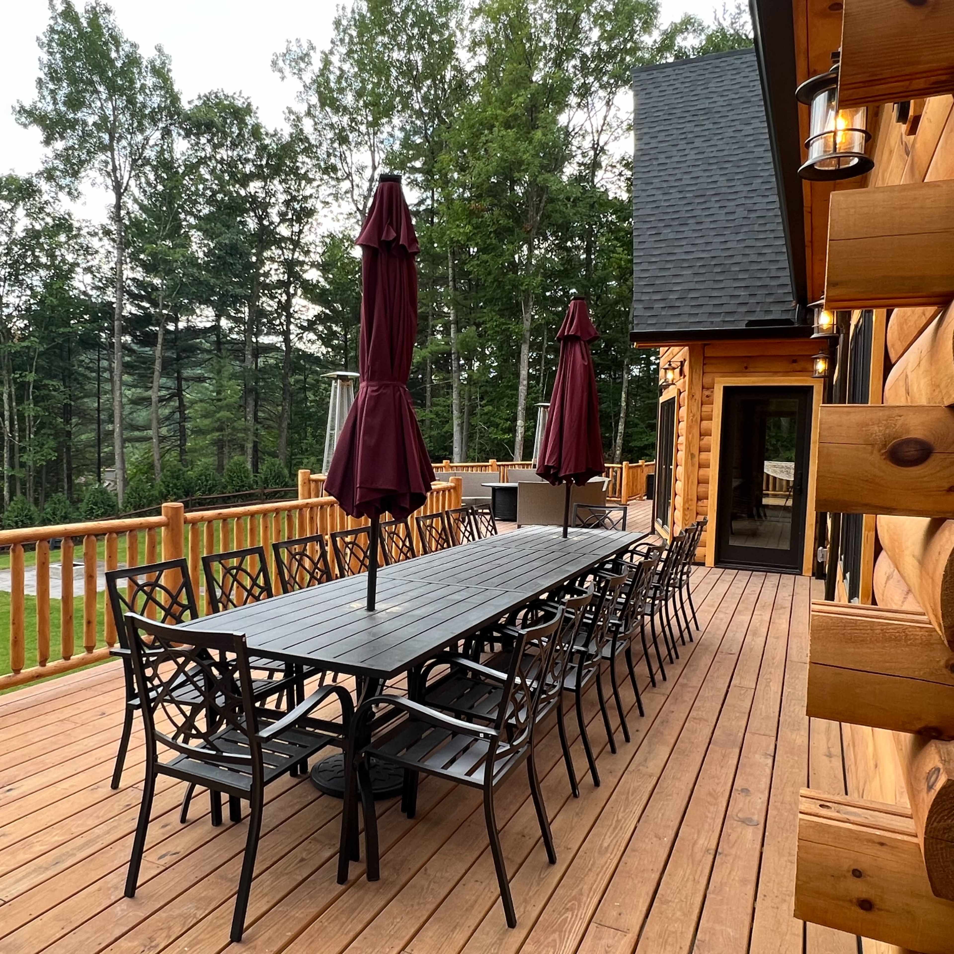 A large wooden deck features a long table surrounded by black chairs, with maroon umbrellas providing shade, overlooking a wooded area.