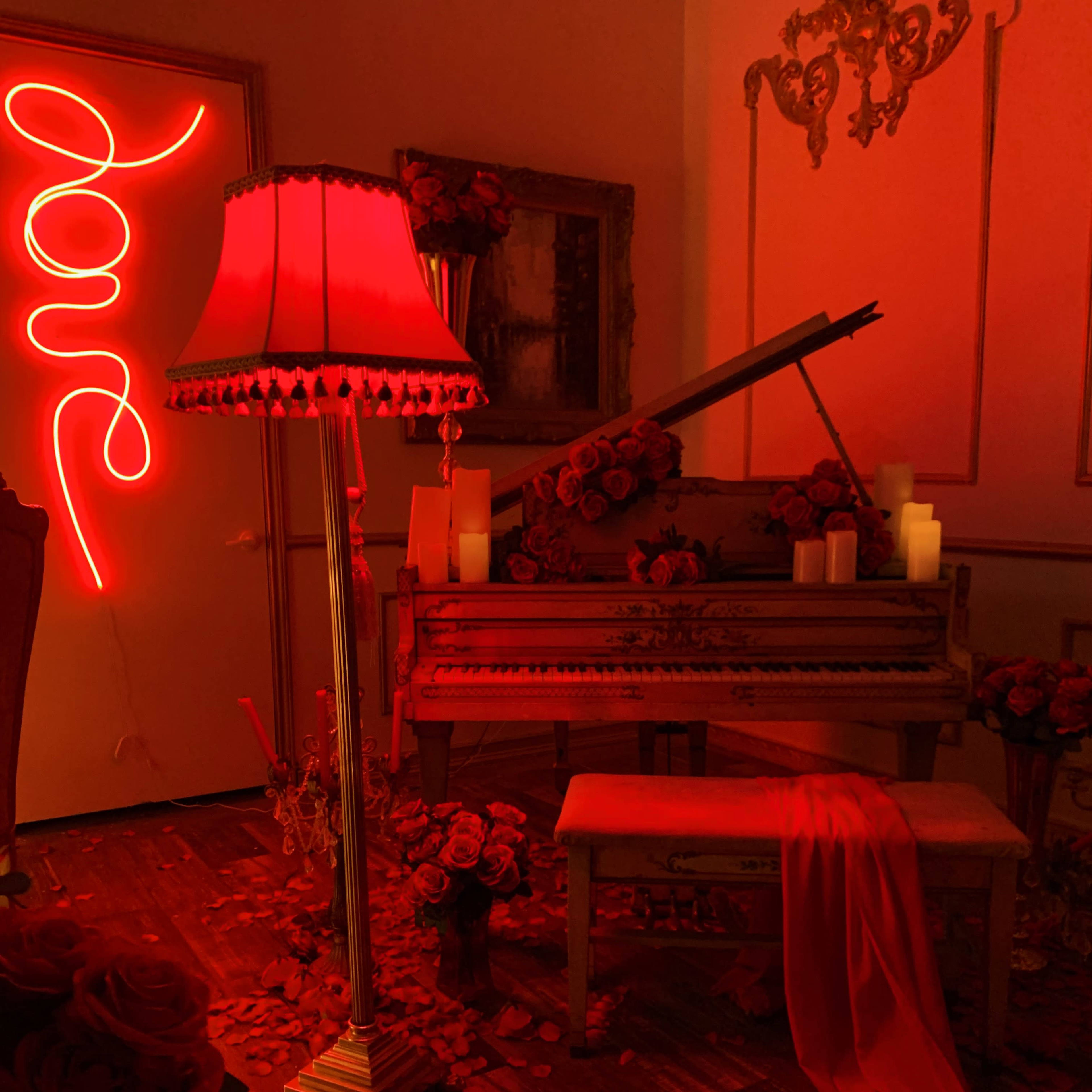 A red-lit room features a vintage piano adorned with roses, candles, and a flowing red fabric.