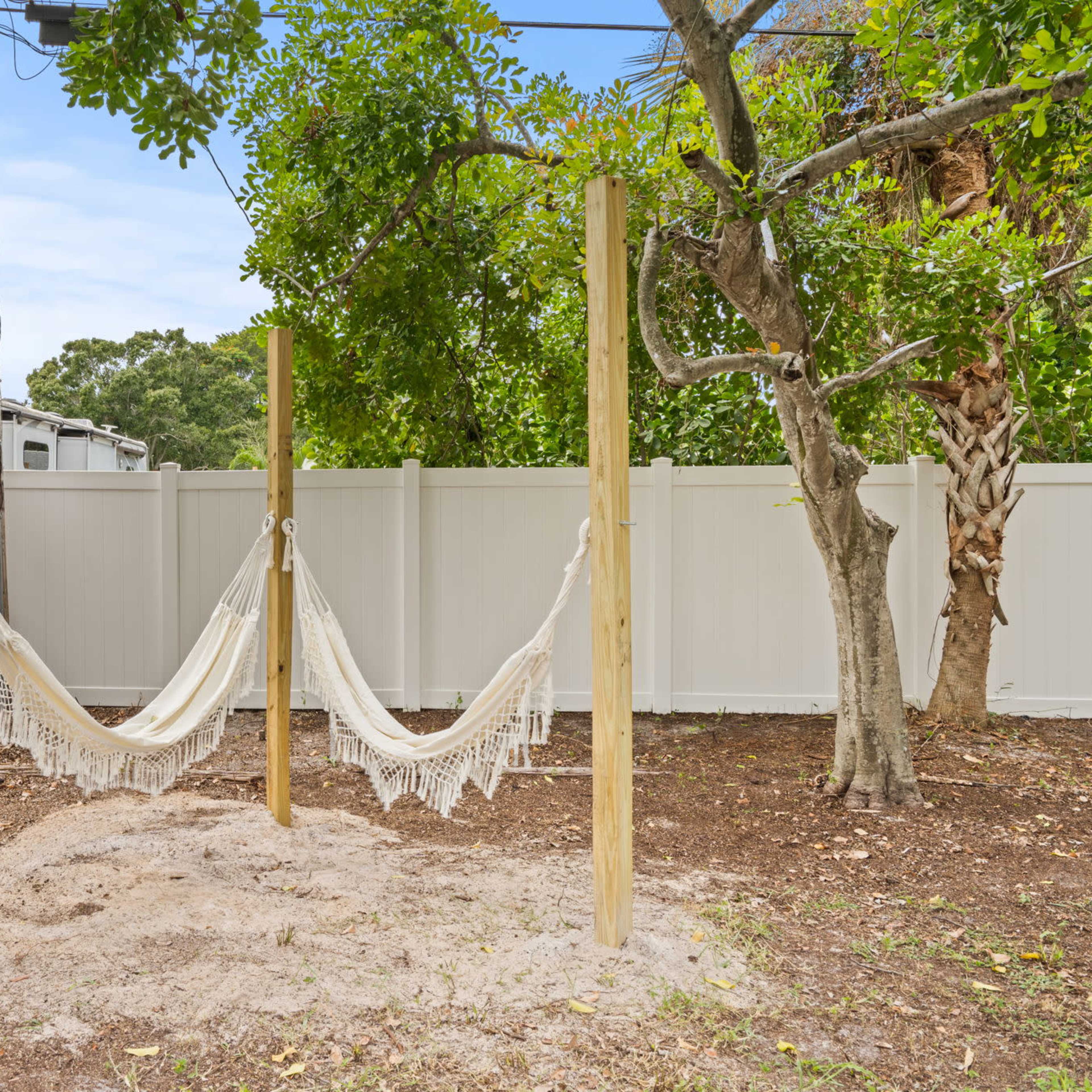 A hammock hung between two wooden posts in a sandy area surrounded by a white fence and trees.
