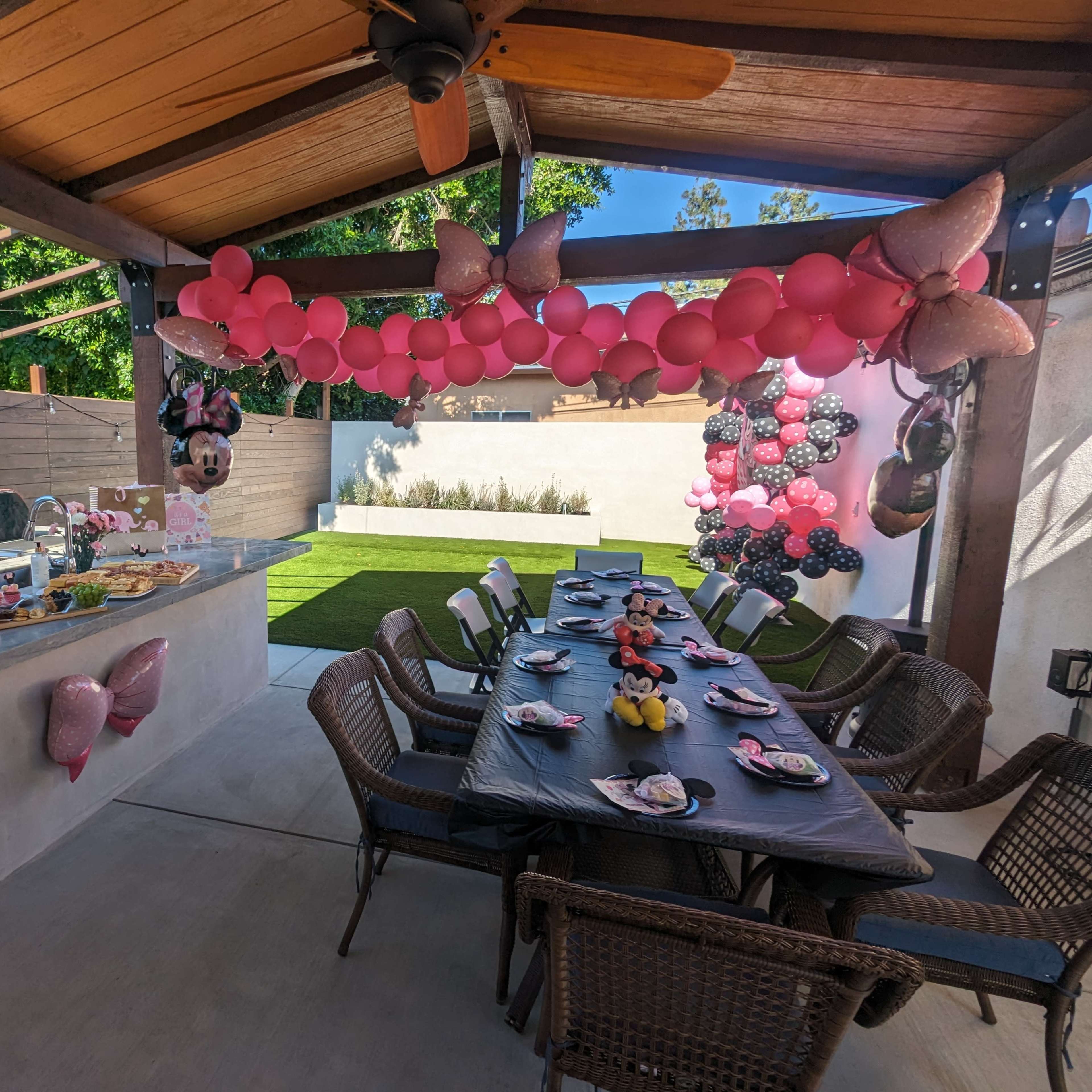 A decorated outdoor dining area features a long table set with plates and party items, surrounded by balloons and a small buffet counter.