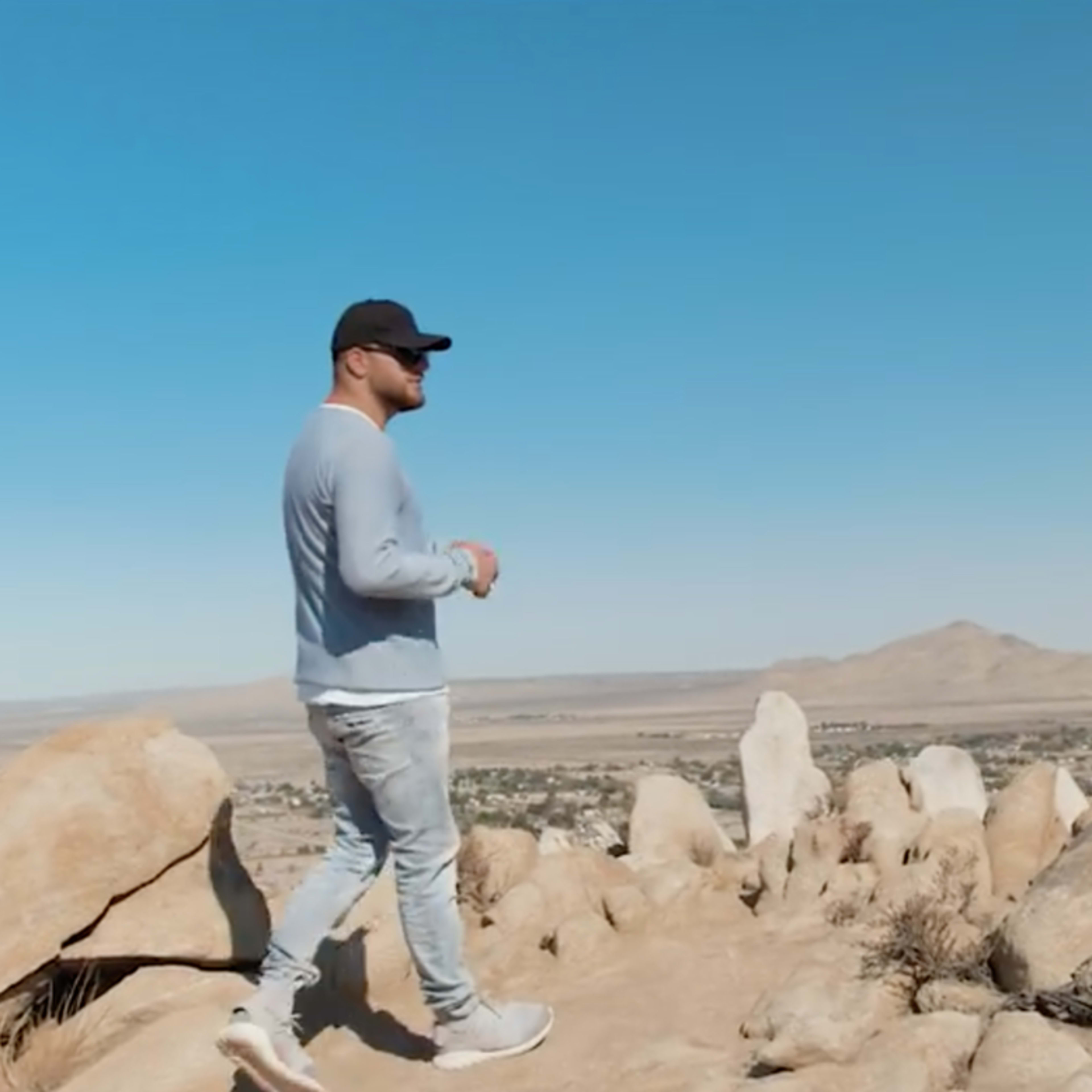 A person is walking on rocky terrain overlooking a wide valley under a clear blue sky.