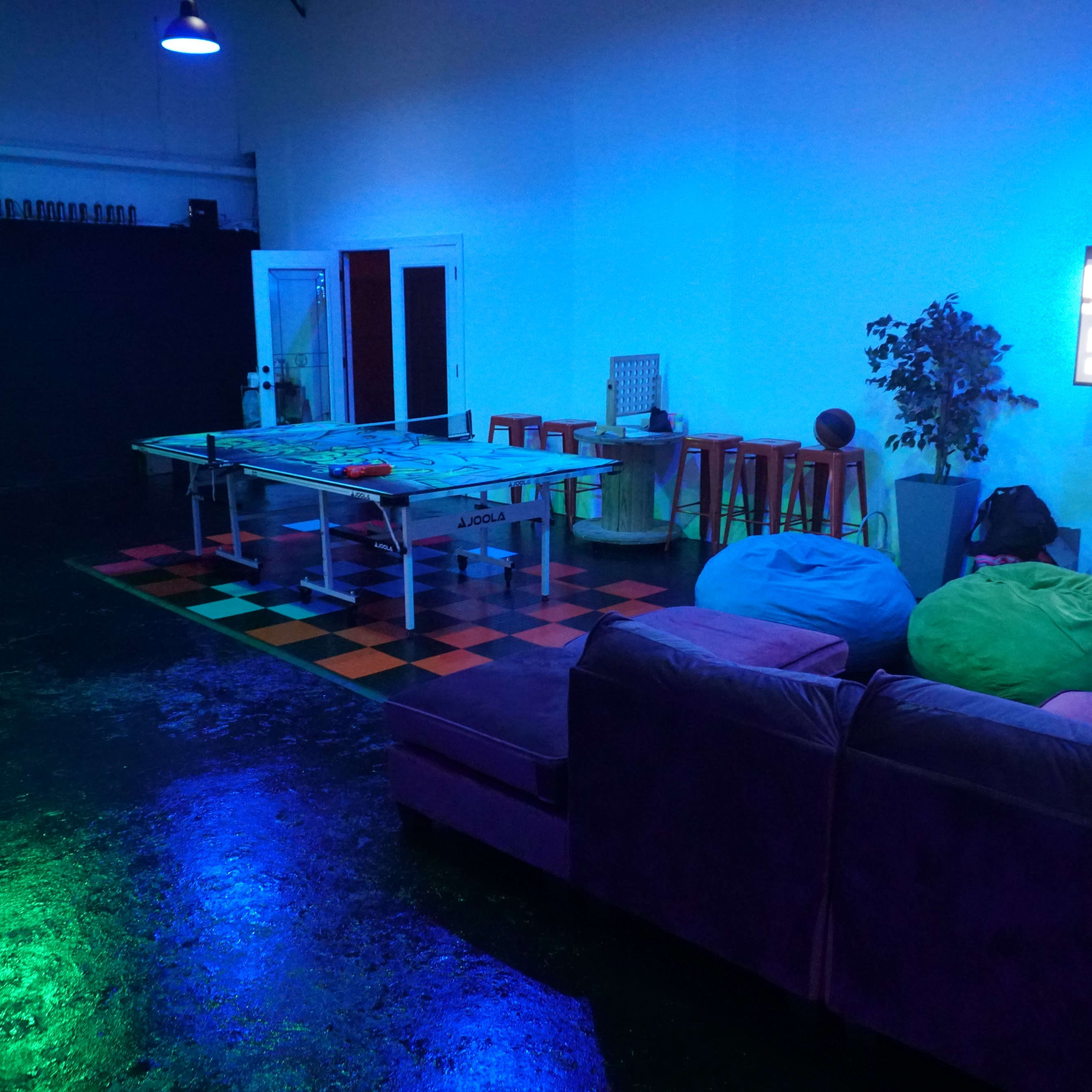 The room features a ping pong table, colorful bean bags, a couch, and a large screen set against a backdrop of blue lighting and black walls.