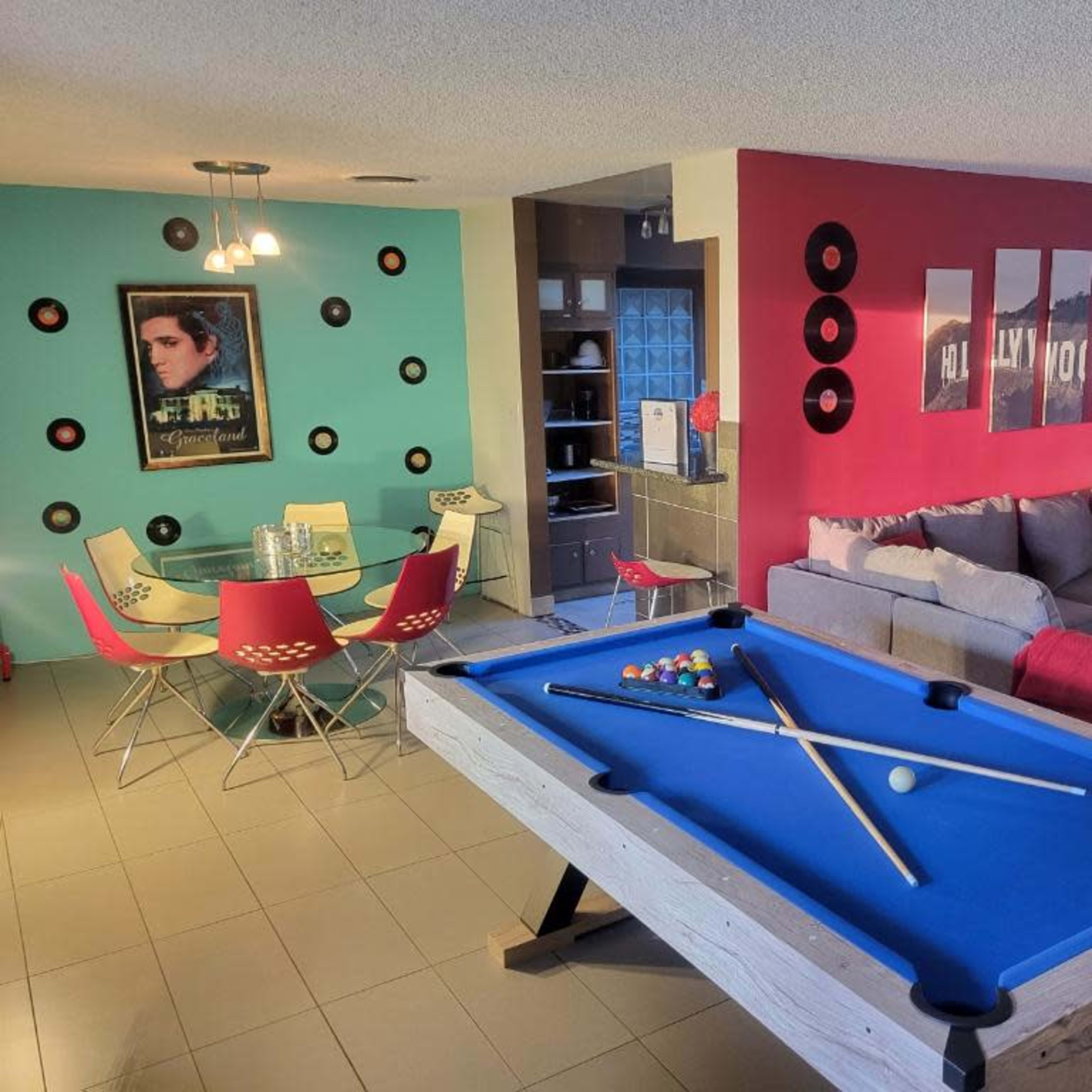 A vibrant room featuring a pool table in the foreground, with a dining area and living space in the background, decorated with colorful artwork and furniture.