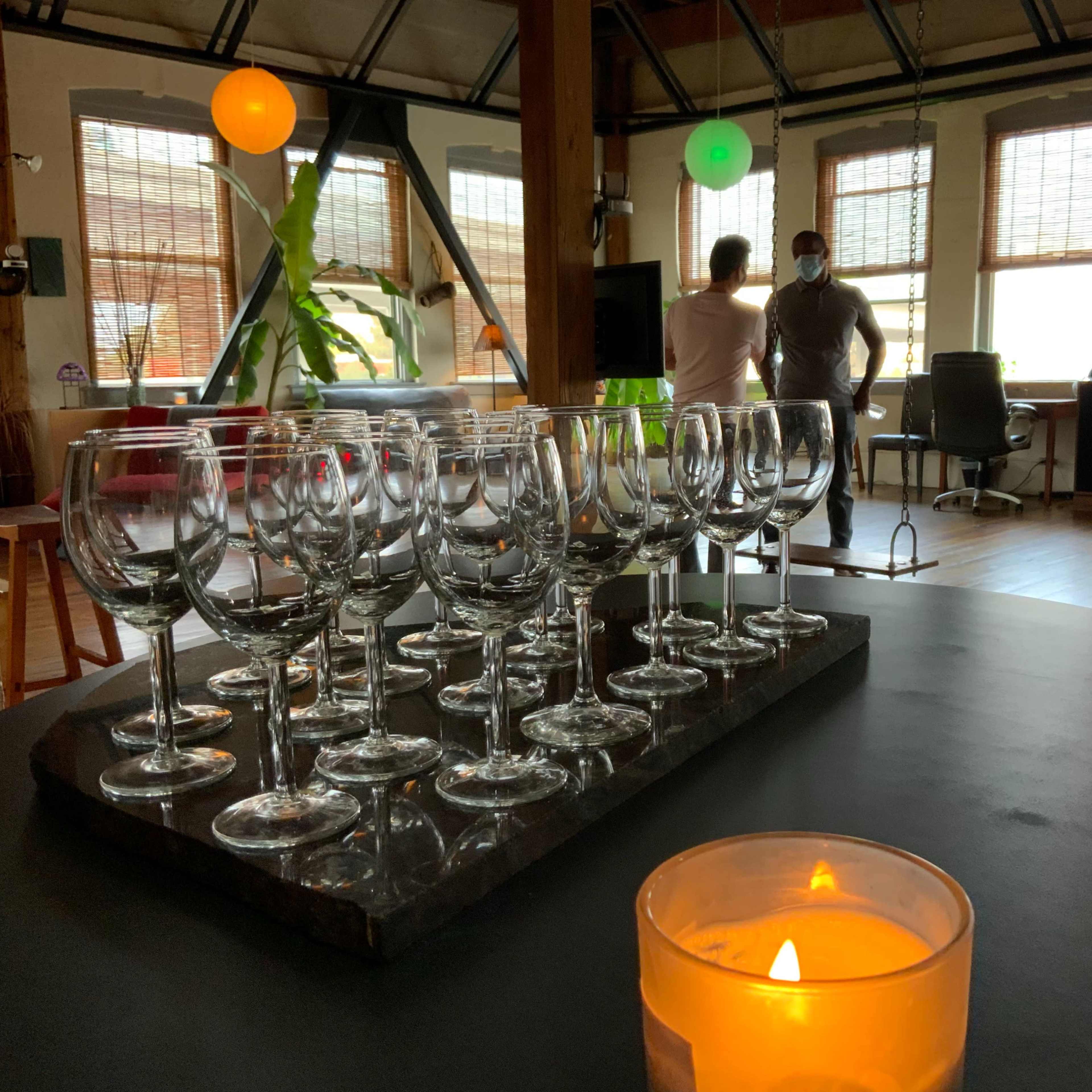 A table with a row of wine glasses in the foreground, while two people converse in a well-lit room decorated with hanging lanterns and potted plants.
