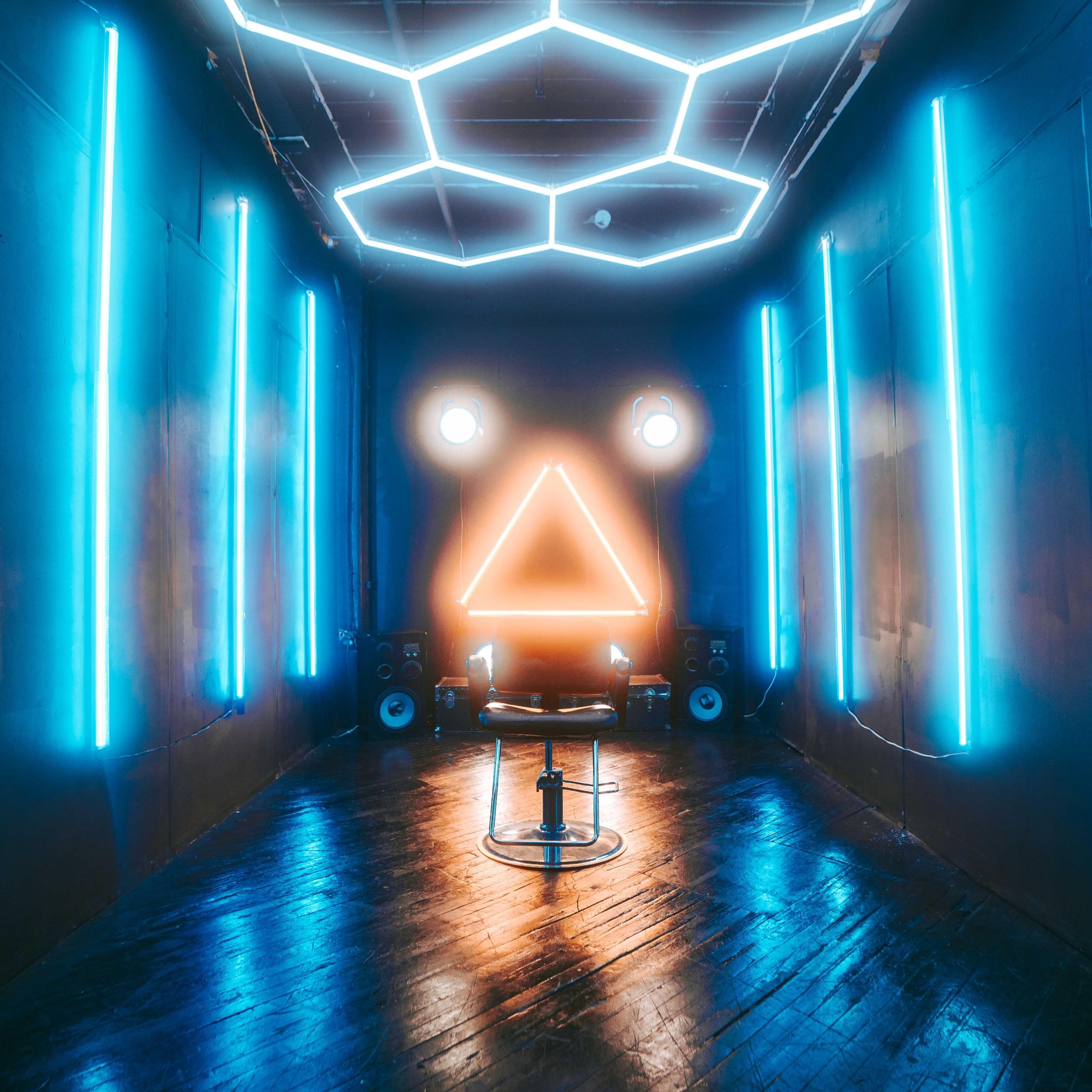 A dimly lit room with glowing blue neon lights arranged in geometric shapes on the walls and ceiling, highlighting a central area with a low seating arrangement.