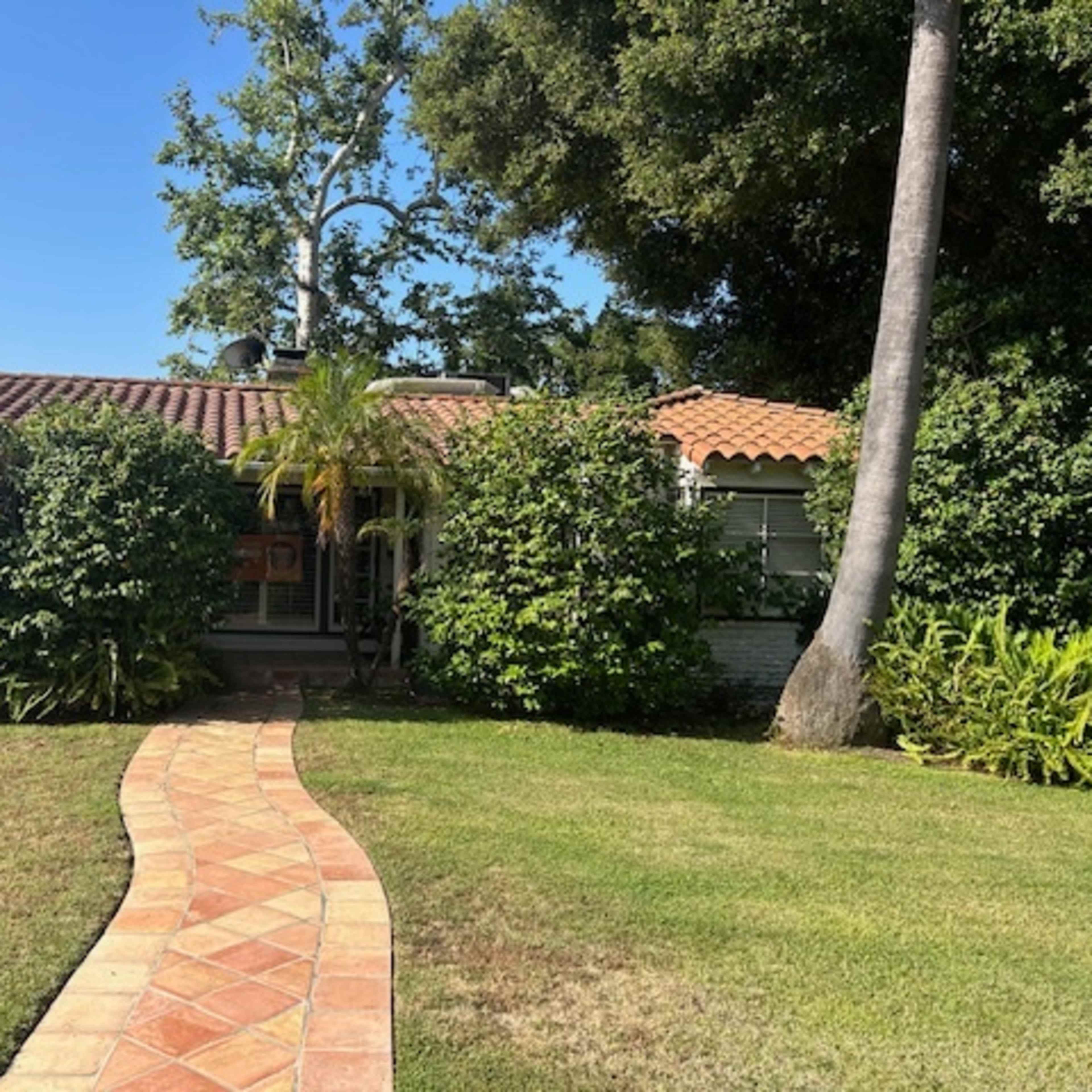 A brick pathway leads through a landscaped yard to a single-story house with a tiled roof and surrounding greenery.