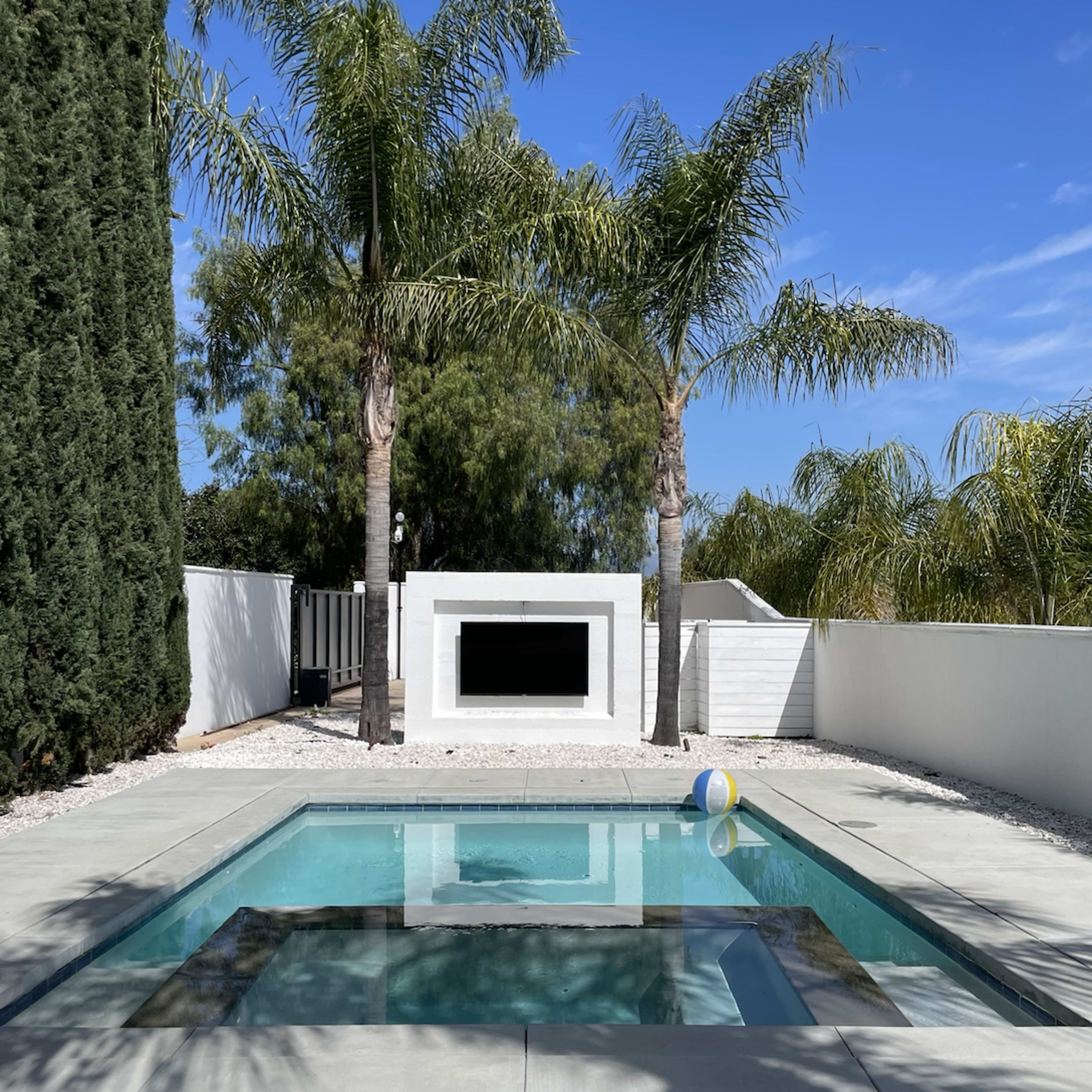 A modern pool area surrounded by palm trees and a white wall with a rectangular opening.