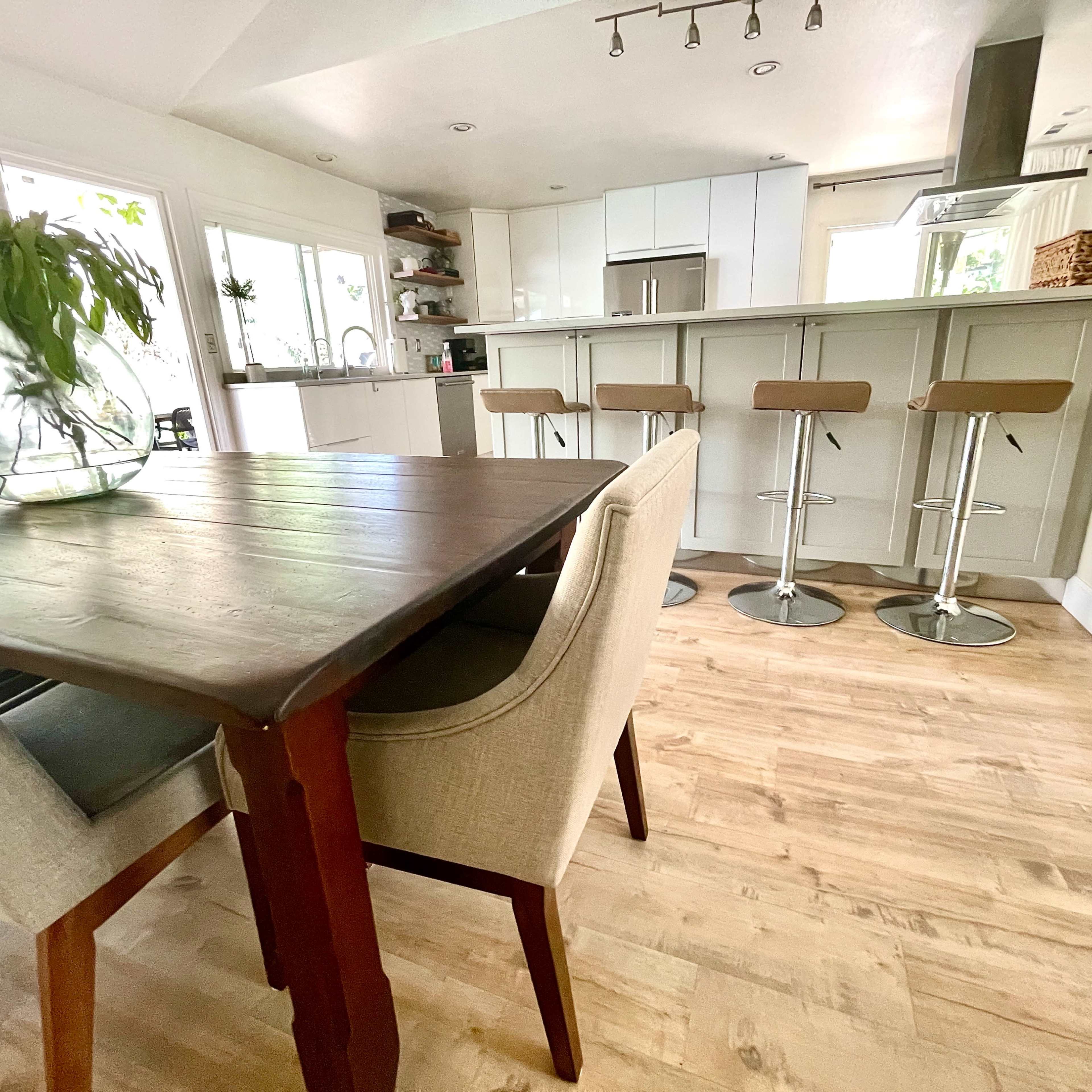 A dining area with a wooden table and upholstered chairs opens into a modern kitchen featuring bar stools and a bright, airy layout.
