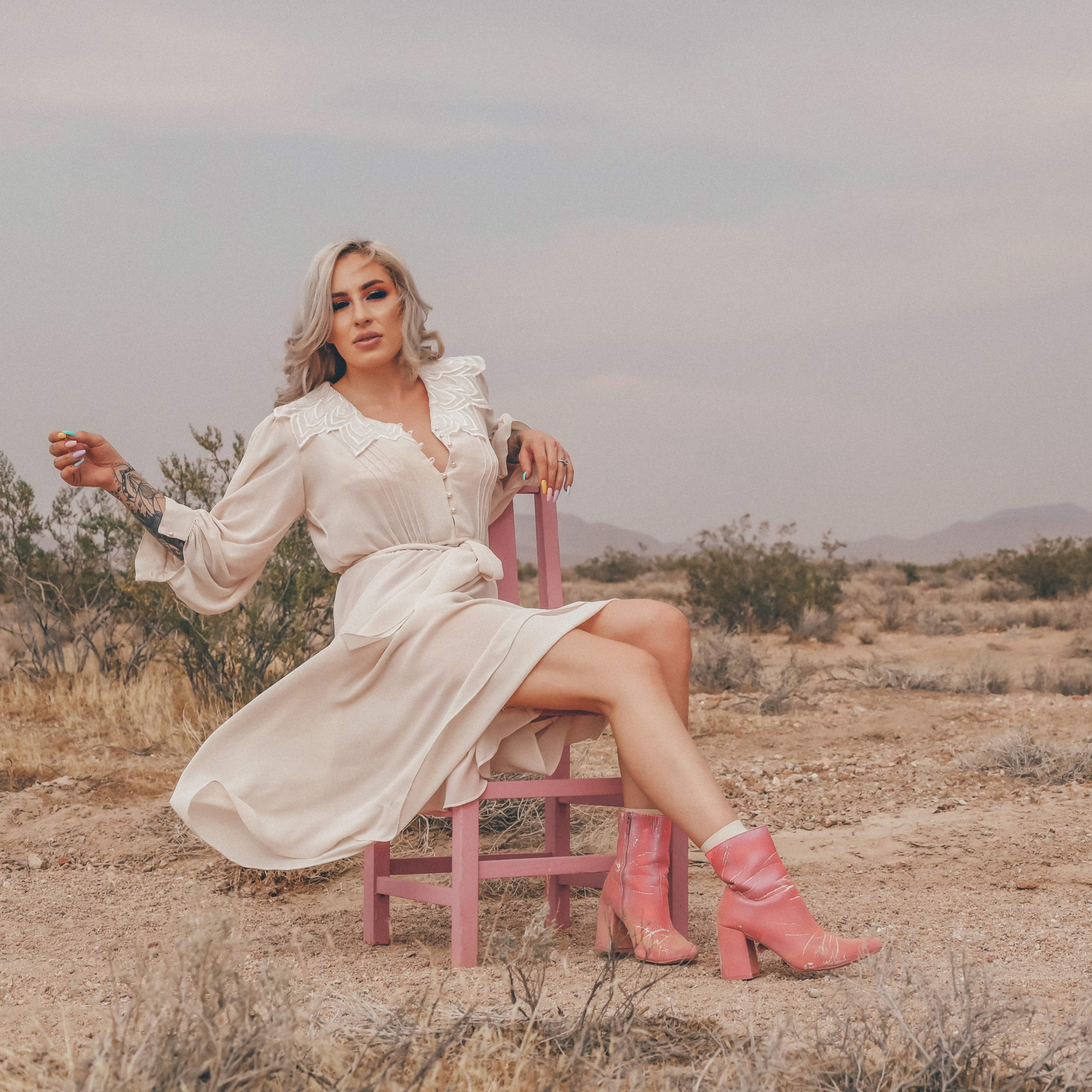A woman in a white dress and pink boots sits on a pink chair in a desert landscape with mountains in the background.