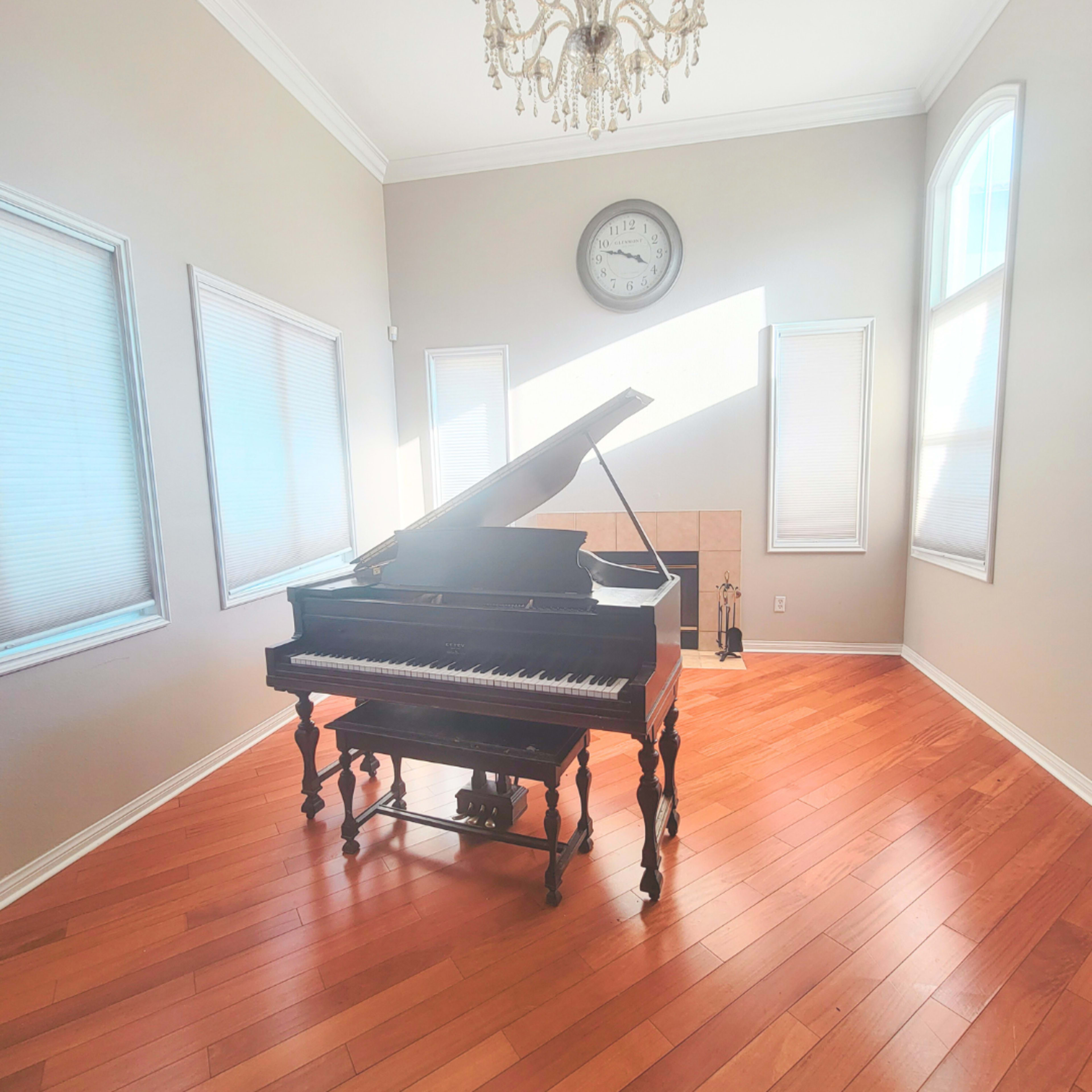 A grand piano is positioned in a room with hardwood floors, large windows, and a chandelier overhead.