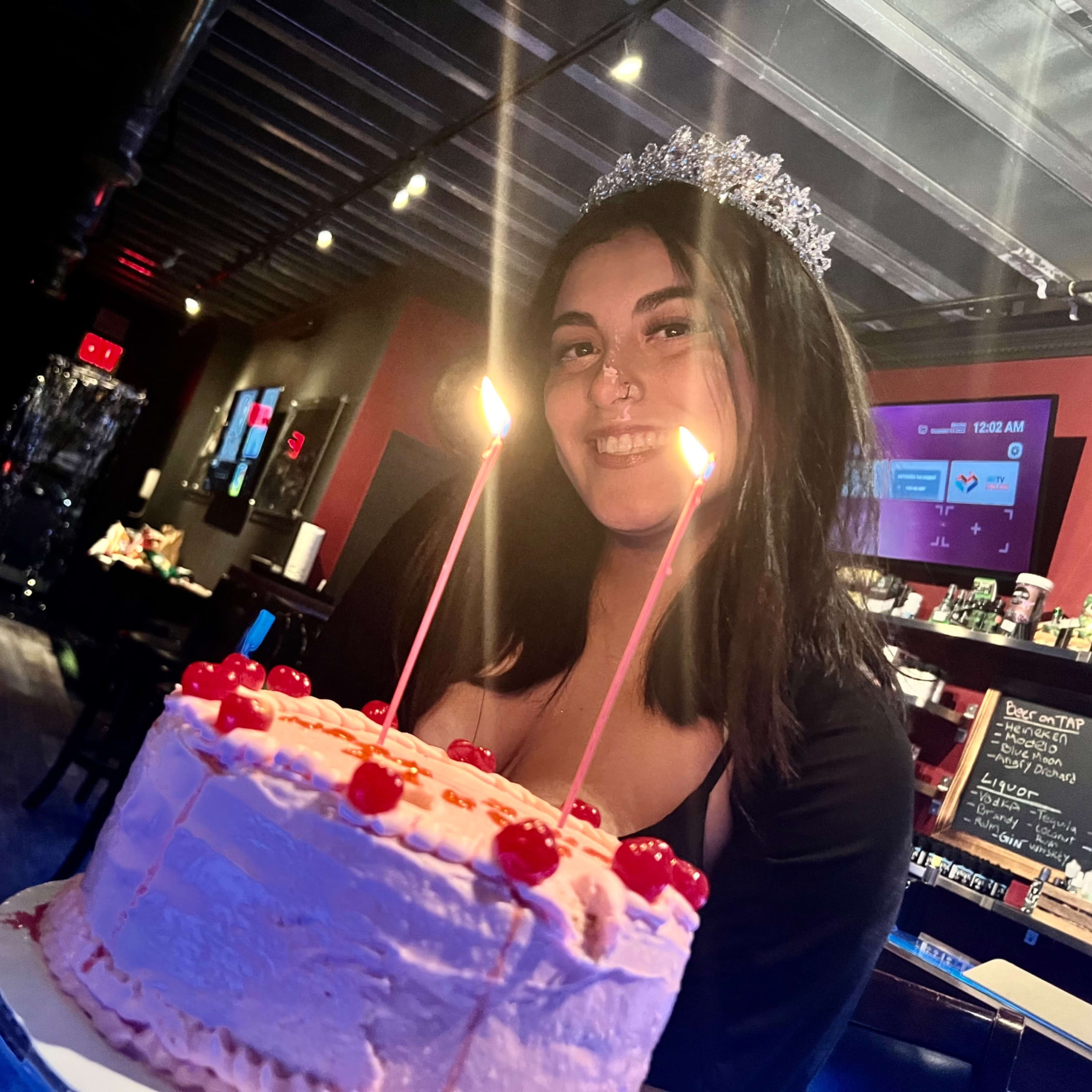 A young woman smiles while holding a decorated birthday cake with candles and cherries on top in a dimly lit venue.