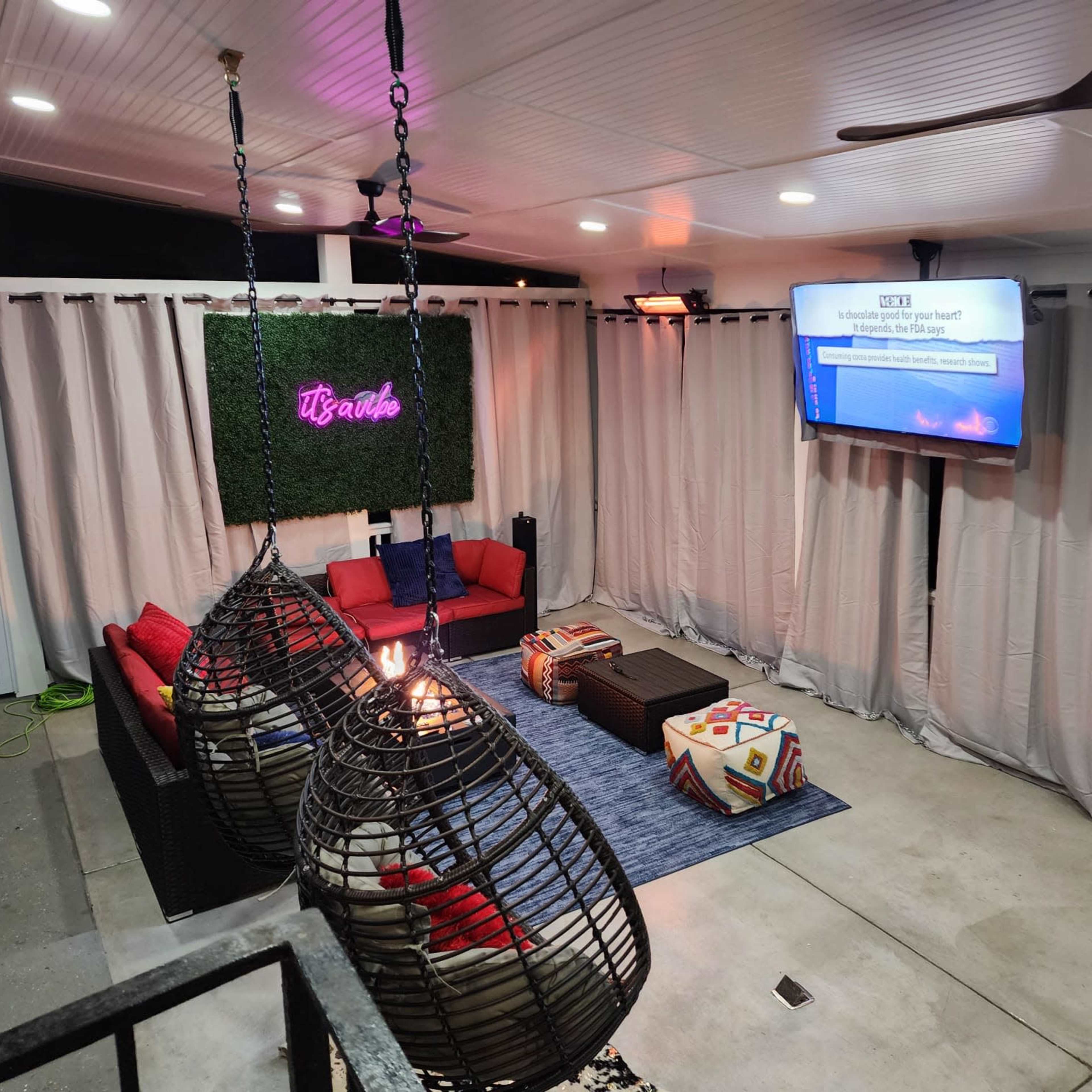 A modern outdoor lounge area with hanging chairs, a television on the wall, and a green plant backdrop.