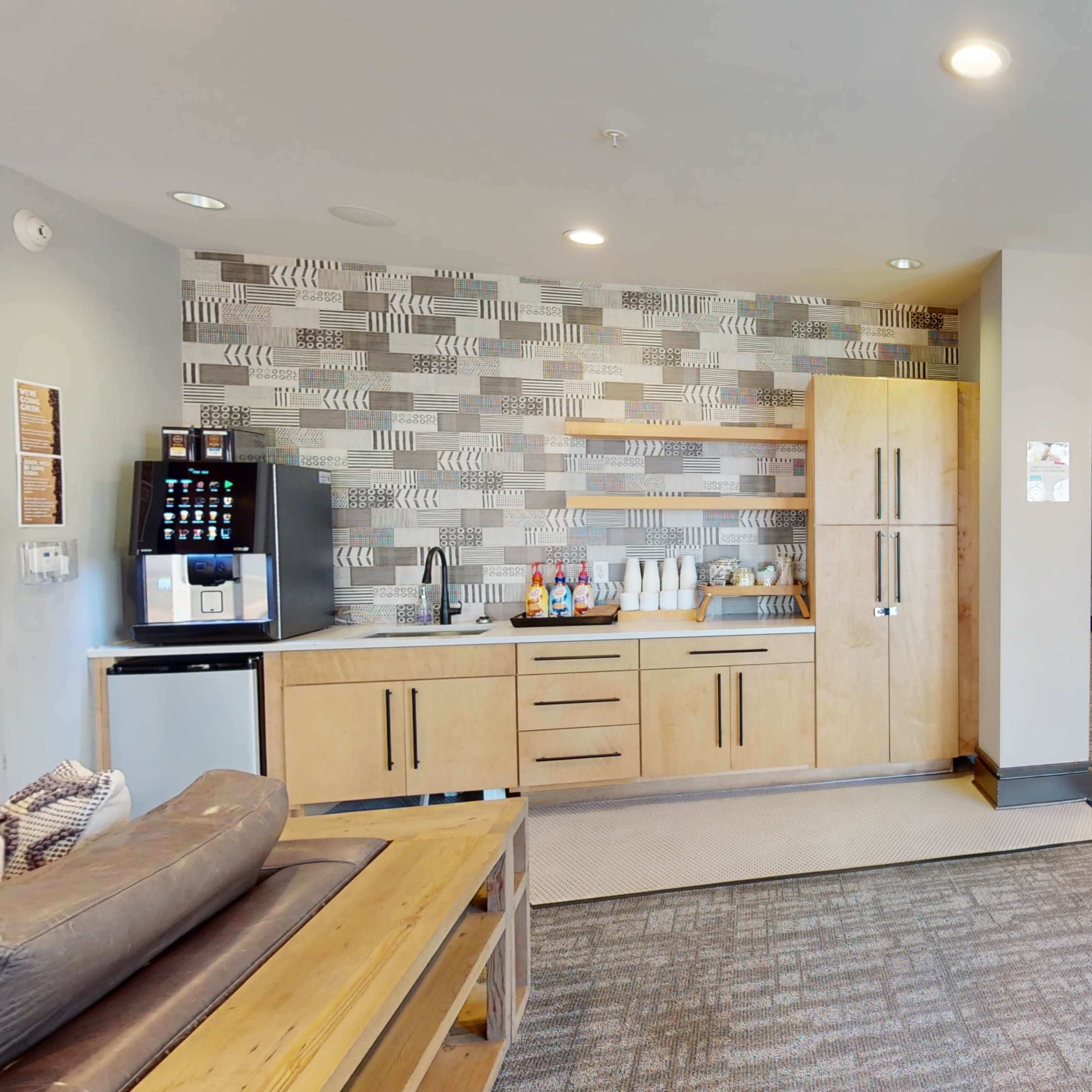 A modern common area with a kitchenette featuring wooden cabinetry, a coffee station, and seating arrangements.