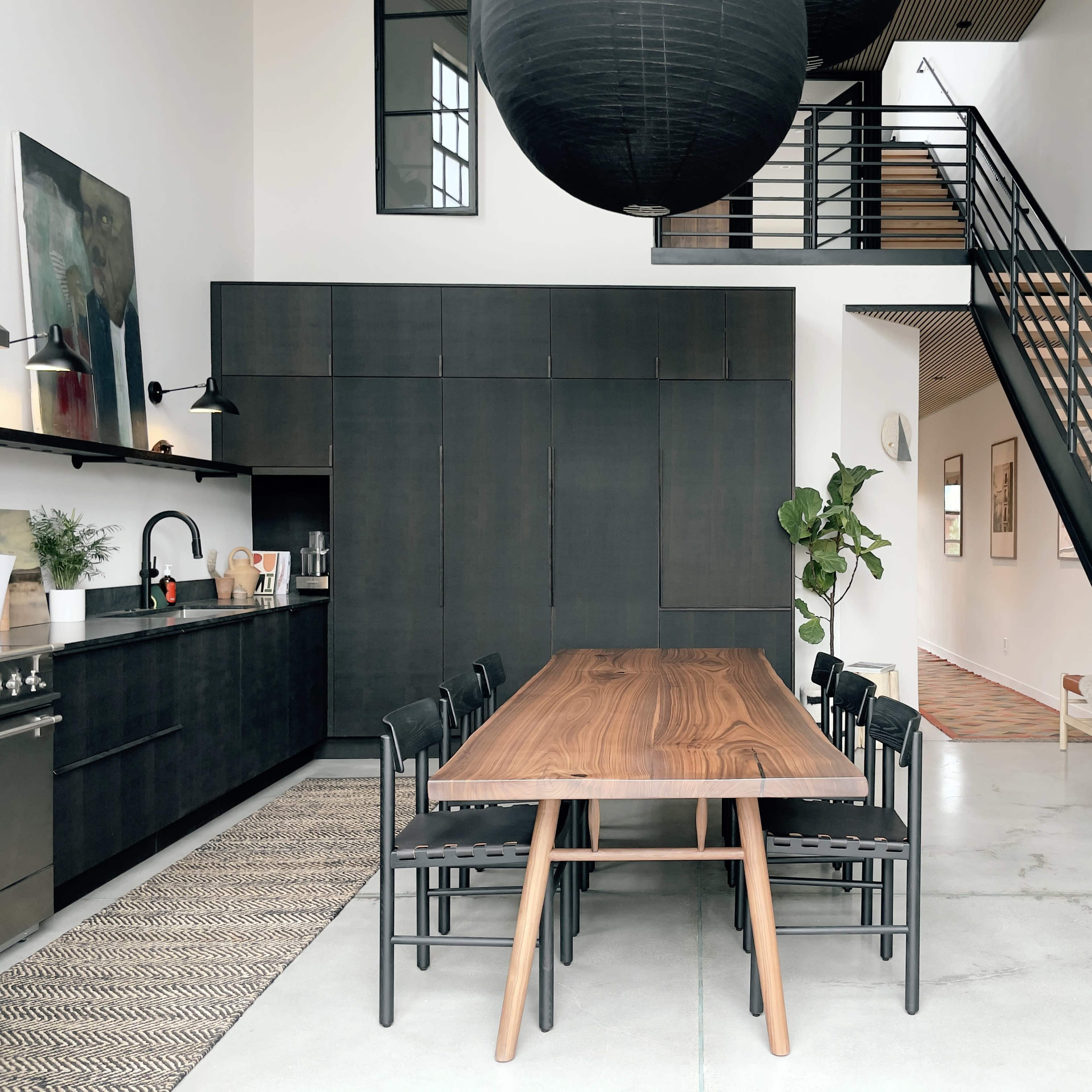 A sleek, modern kitchen and dining area with a large wooden table, black cabinetry, and a staircase leading to an upper level.