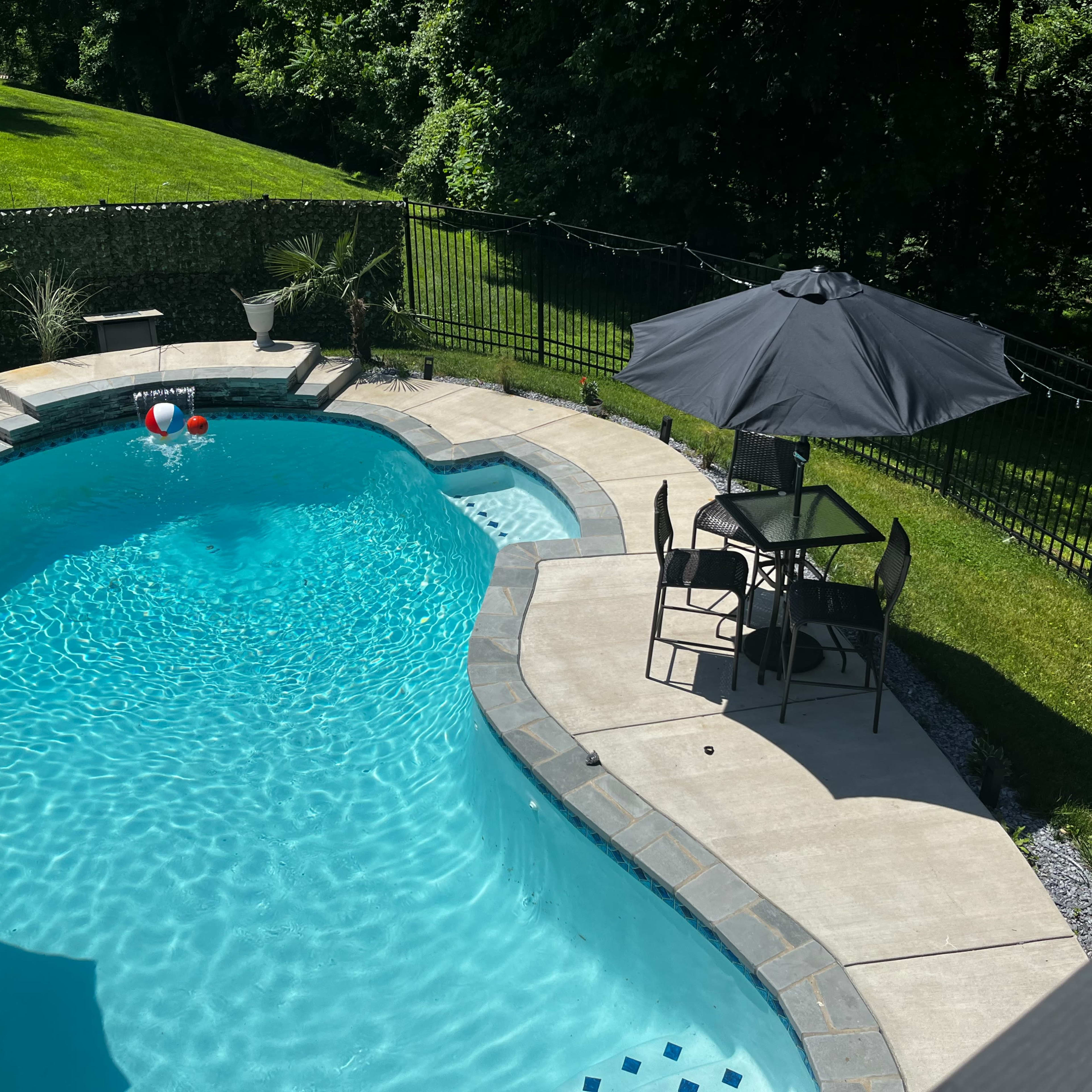 A backyard swimming pool surrounded by a concrete patio, lawn, and a table with chairs under a black umbrella.