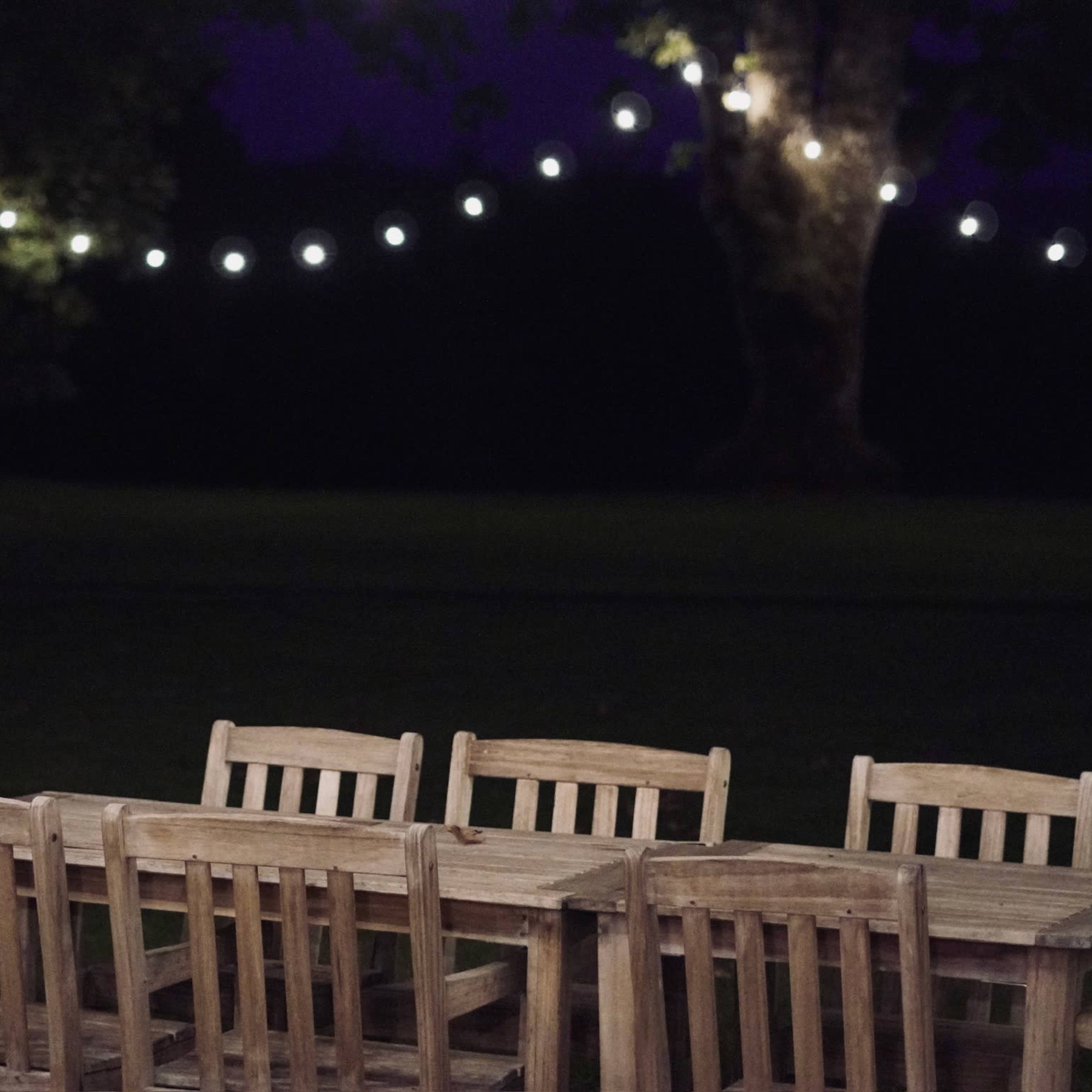 A wooden dining table with matching chairs is set up outdoors under string lights against a dark background.