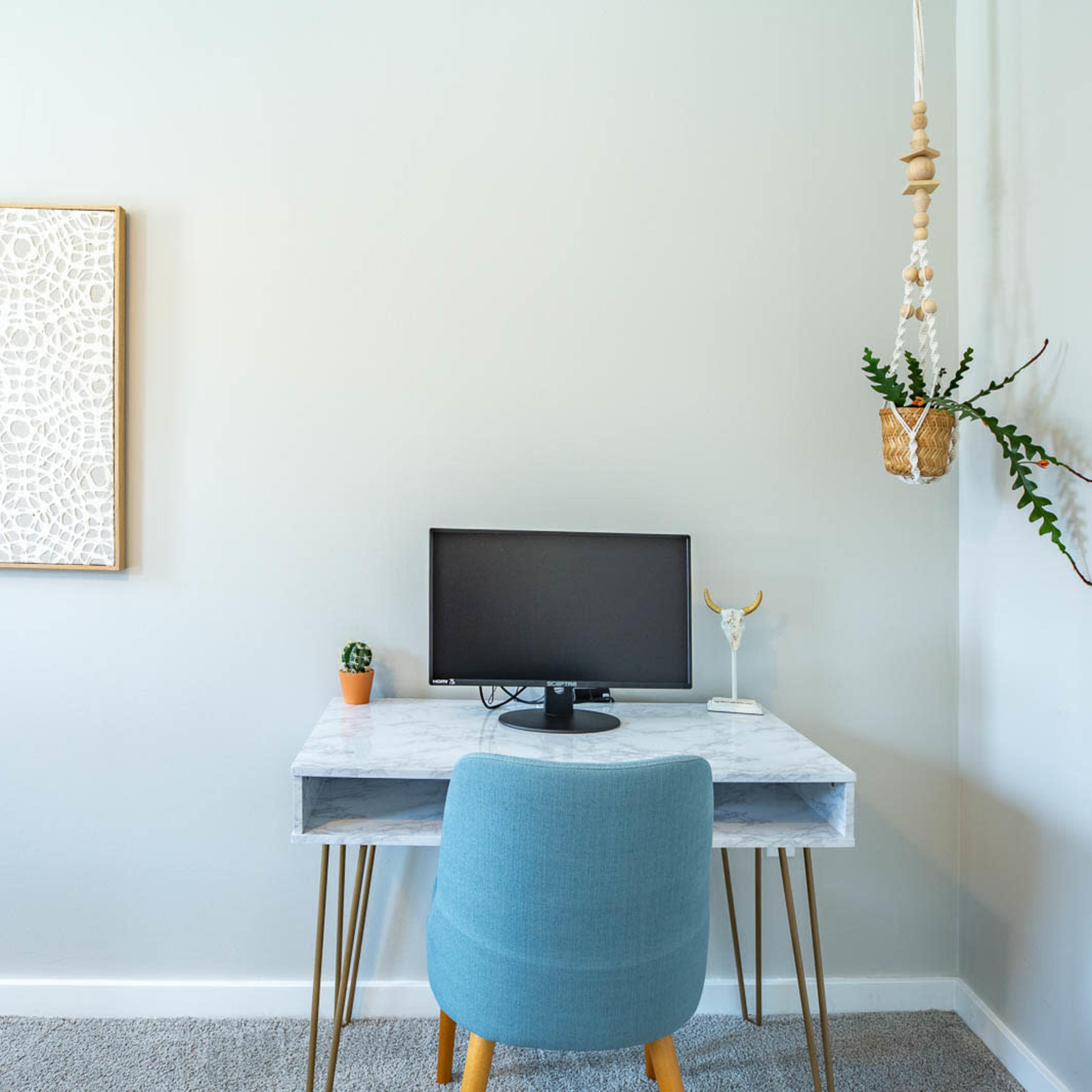 A minimalist workspace features a light-colored desk with a computer monitor, a small plant, and a hanging planter on the wall.