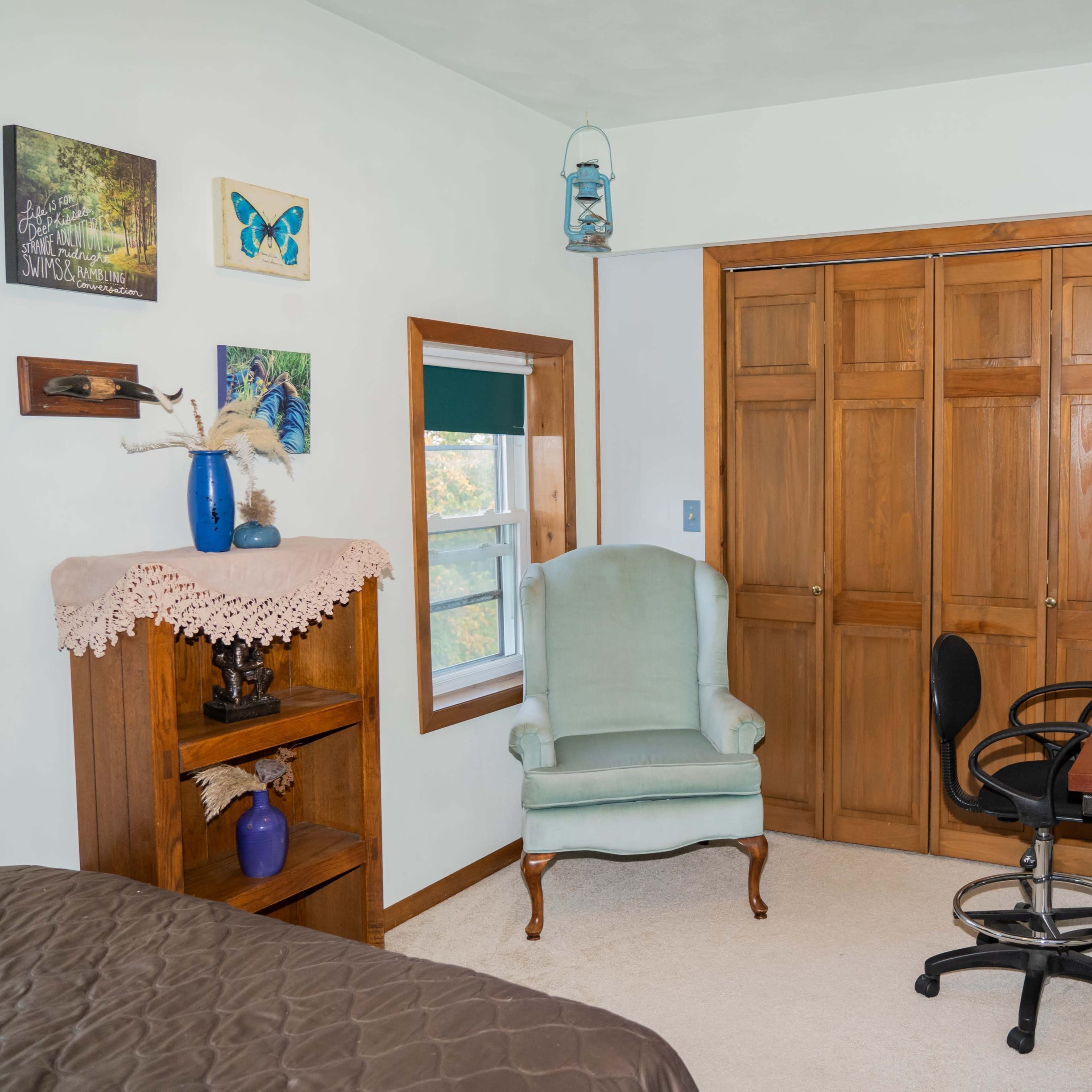 A small room features a green armchair, a wooden bookshelf, a desk with a black chair, and a closet with double doors.