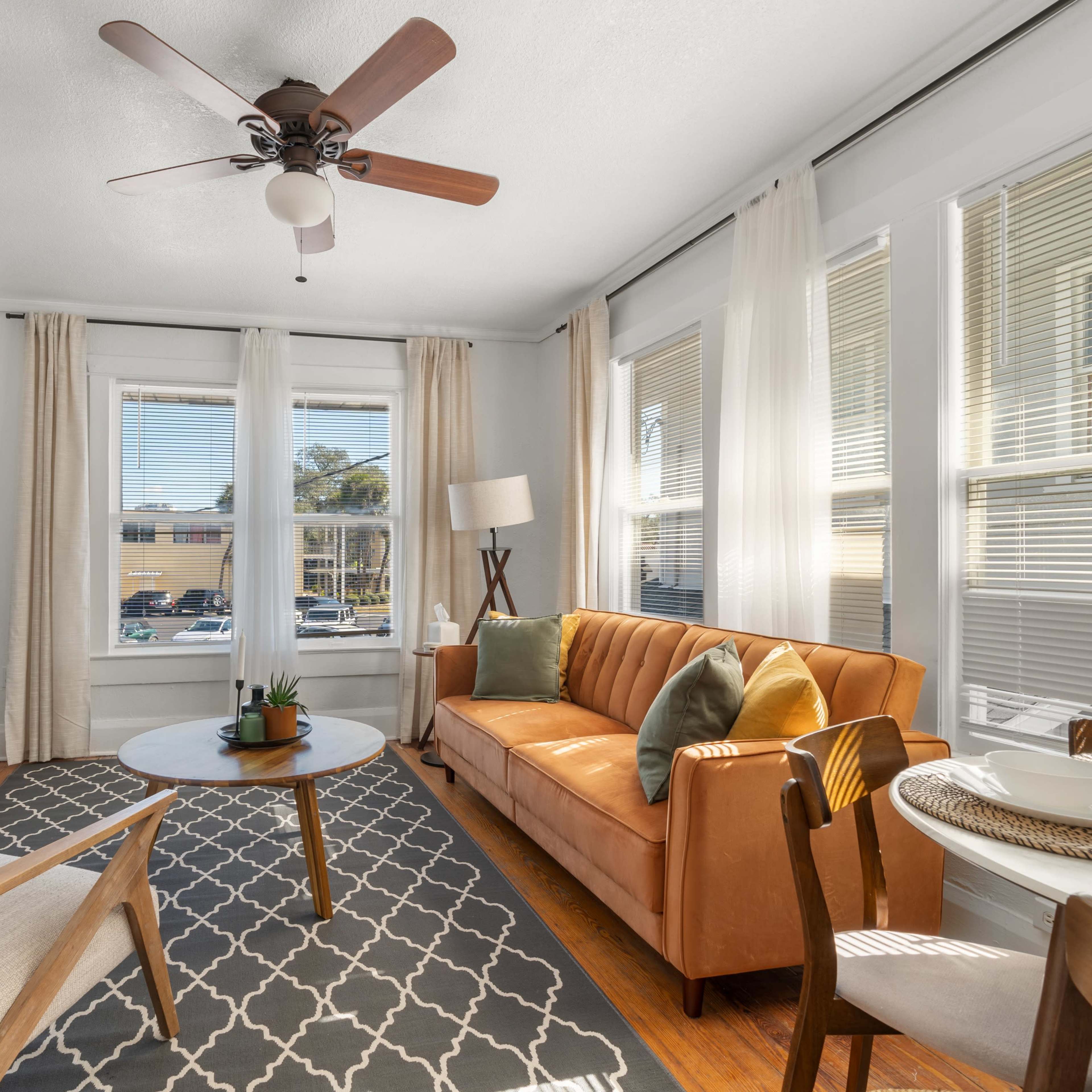 The living room features a tan sofa, light wood furniture, and large windows with sheer curtains, creating a bright and cozy space.