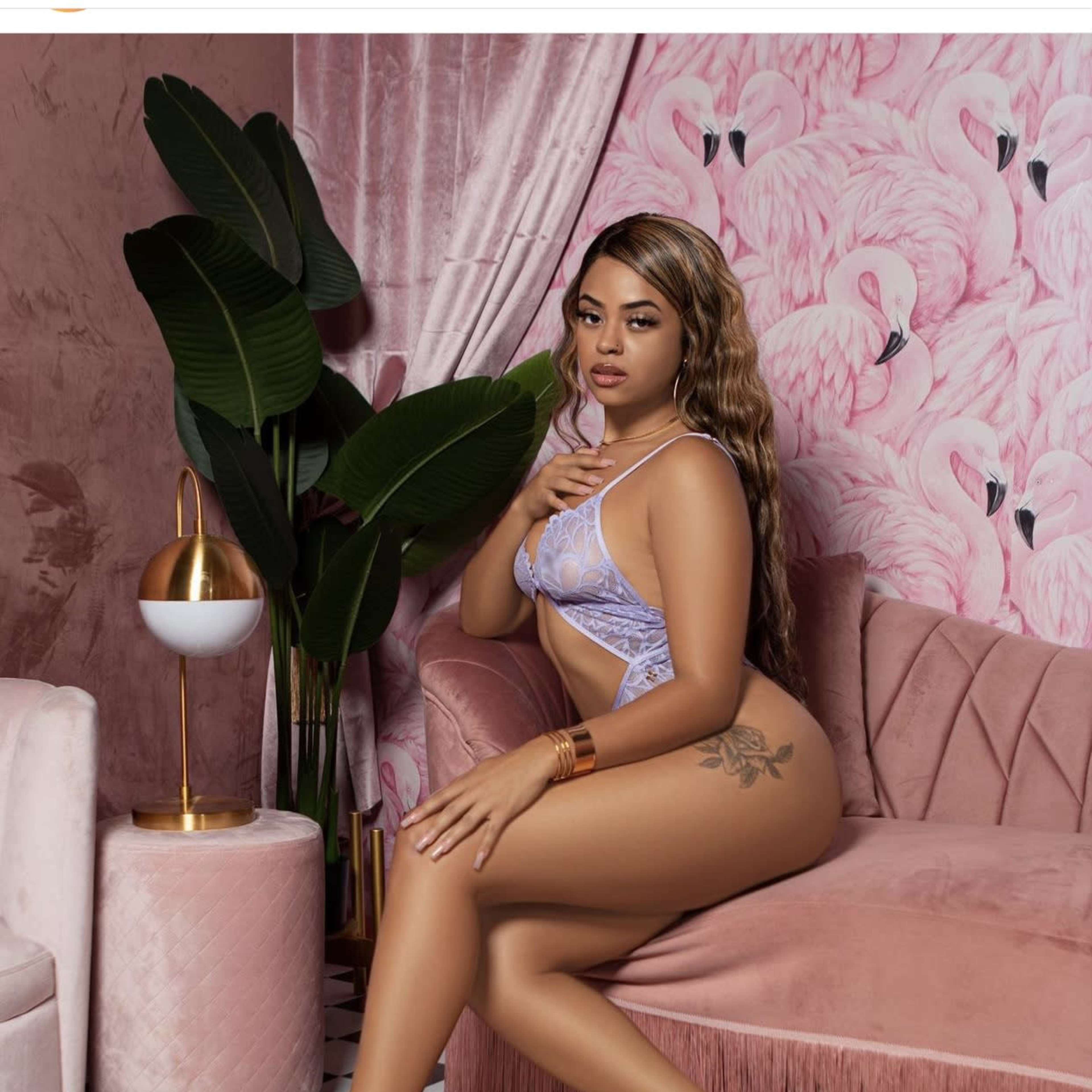 A woman in lingerie seated on a pink couch in a room with a flamingo-patterned wall and a decorative plant.