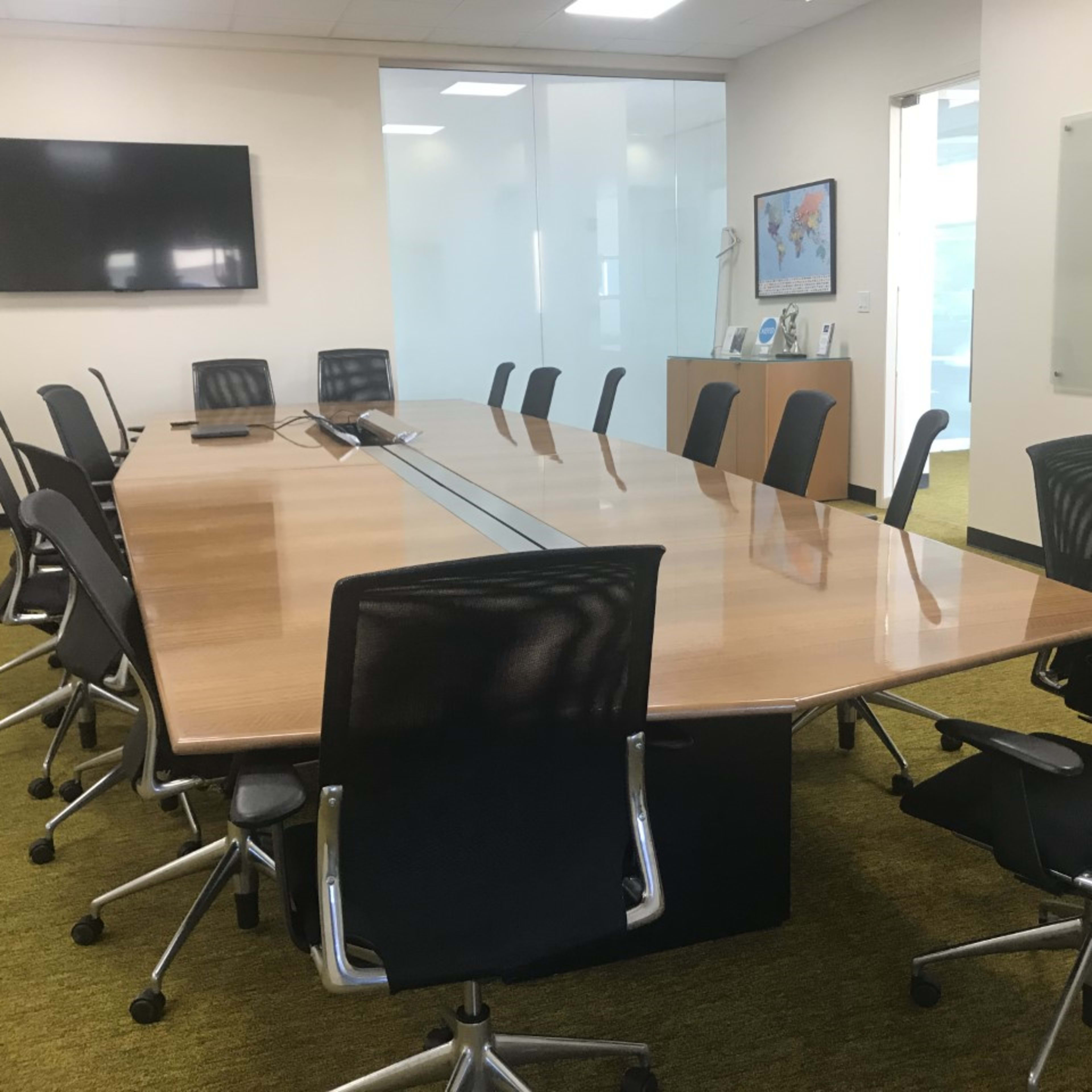 A large conference room features a rectangular wooden table surrounded by ergonomic black chairs, with a wall-mounted screen and a glass partition.
