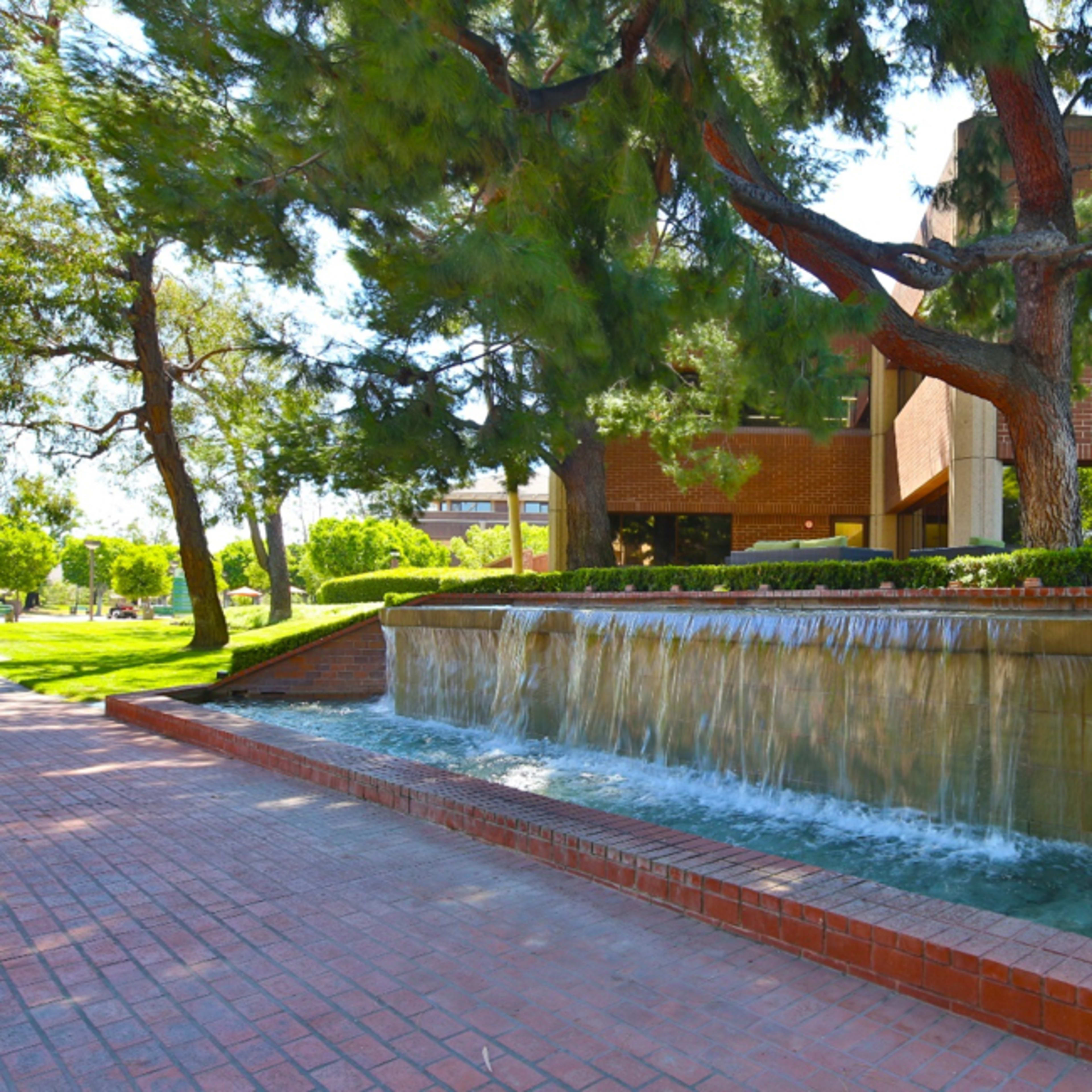 A low stone fountain with cascading water, surrounded by trees and a brick pathway adjacent to a modern building.