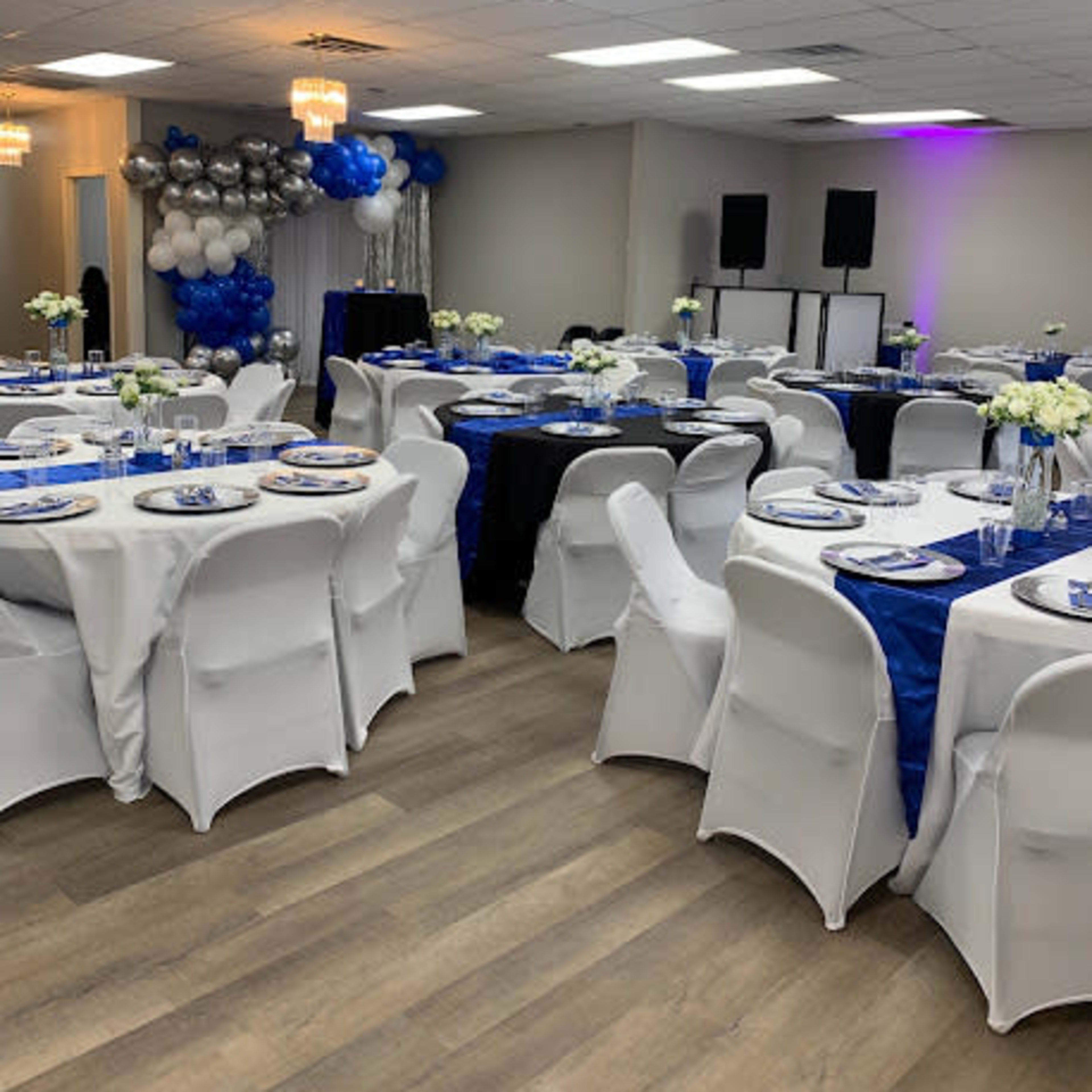 A decorated event space with tables set for dining, featuring white and blue tablecloths, floral centerpieces, and balloon decorations.