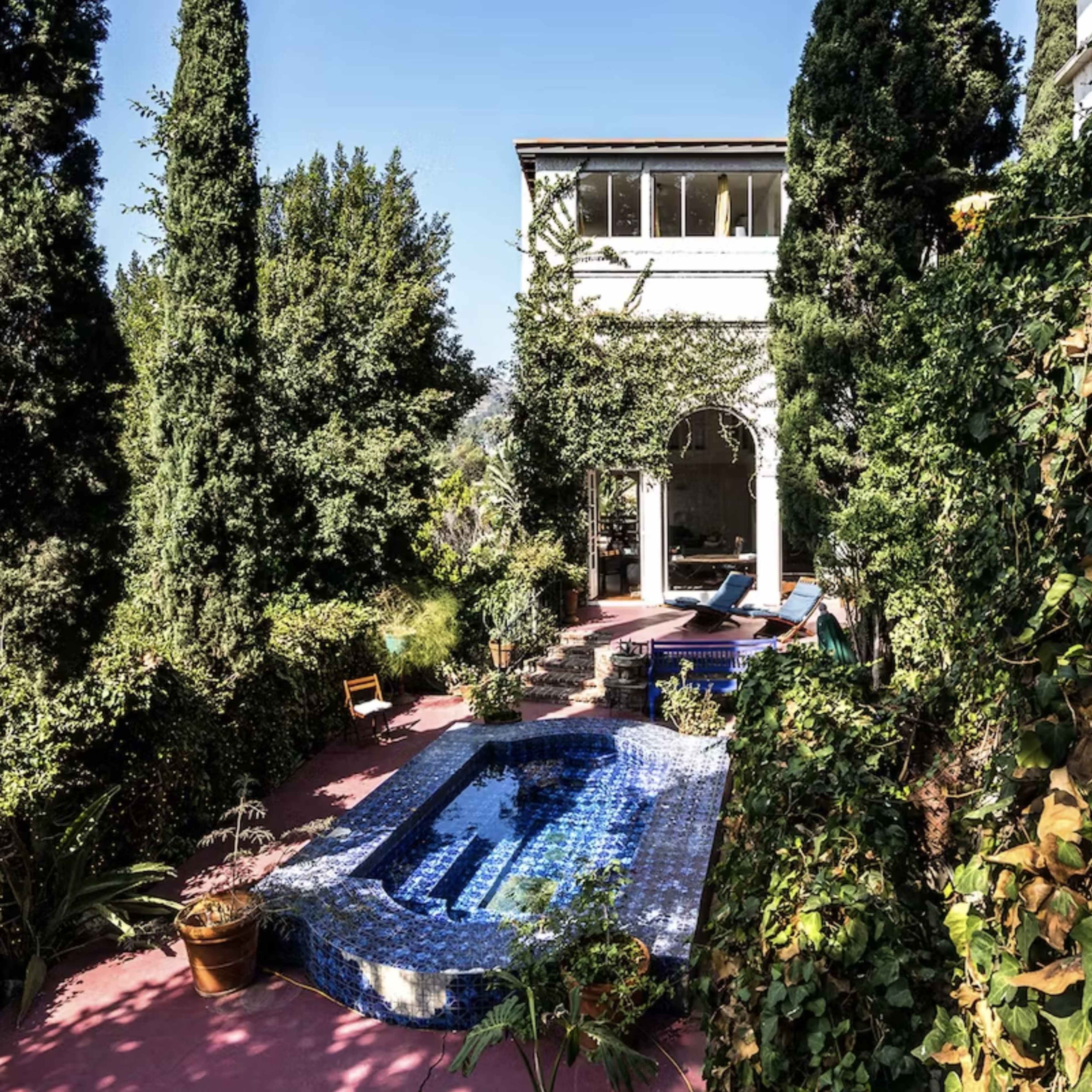 A mosaic-tiled pool is surrounded by lush greenery and tall cypress trees beside a multi-level building.