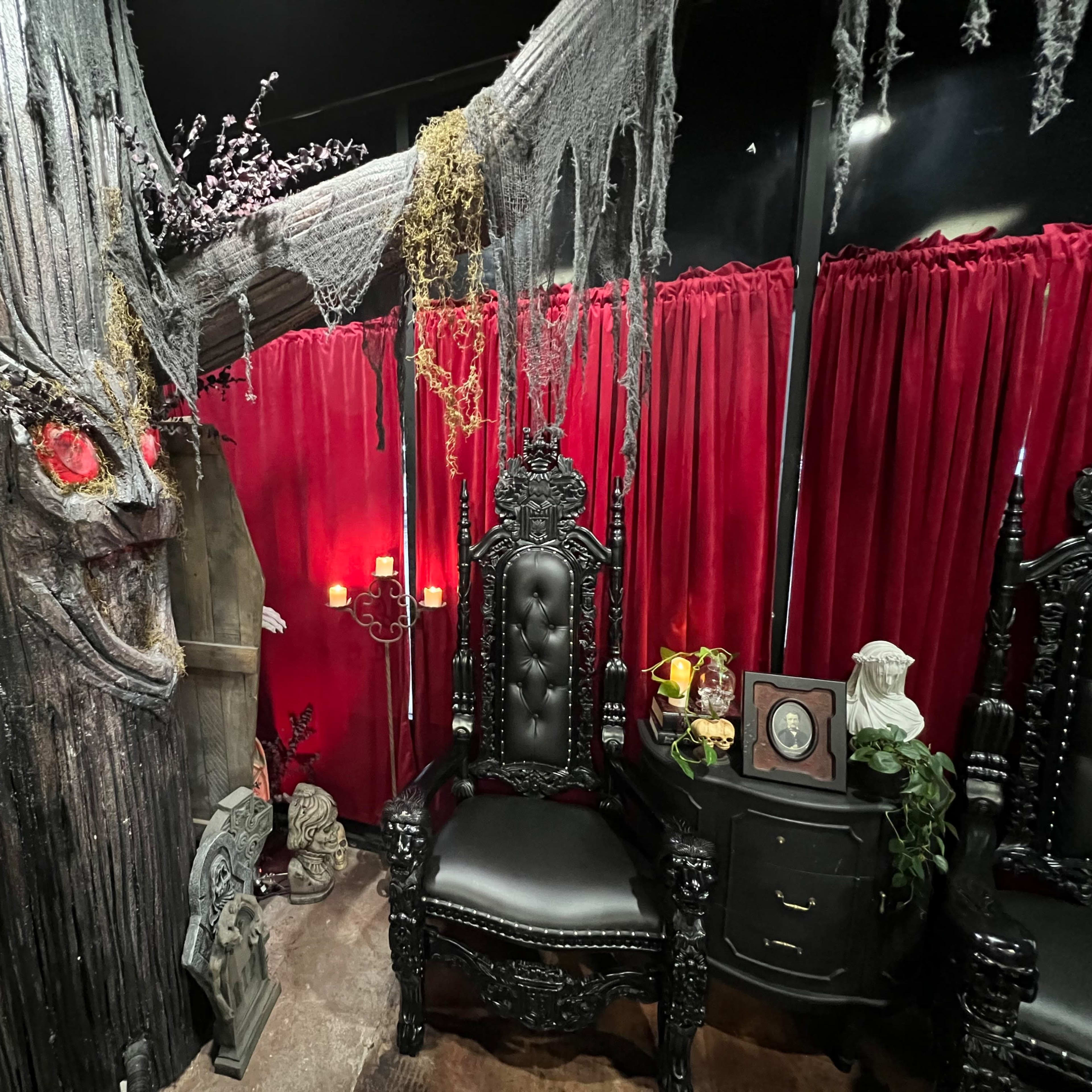 The scene features a dark, elaborate room with a large, gnarled tree, two ornate black chairs, red drapes, and eerie decorations including skulls and skeletons.