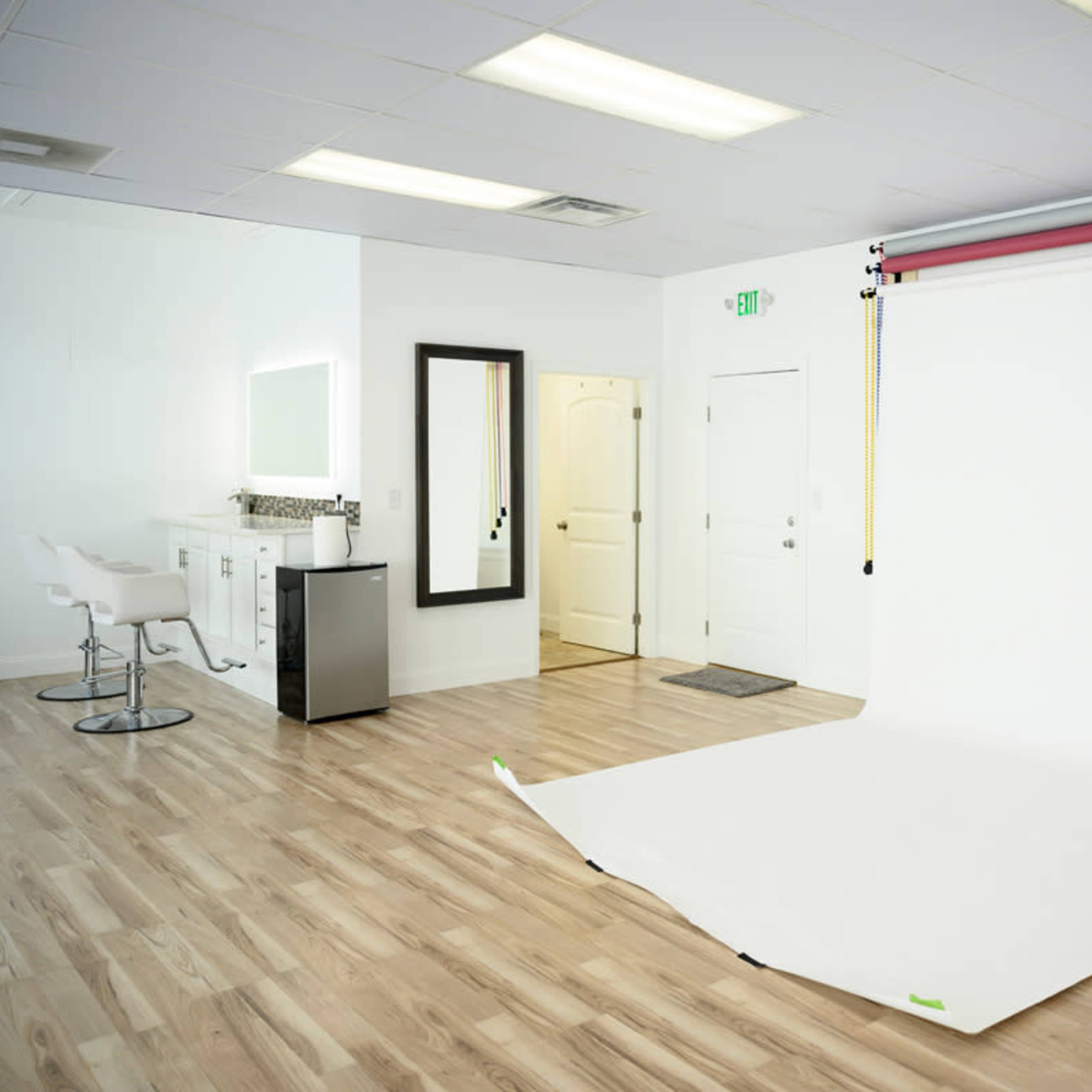 A spacious studio with a white backdrop set up for photography, featuring a mirror, a small counter, and modern seating.