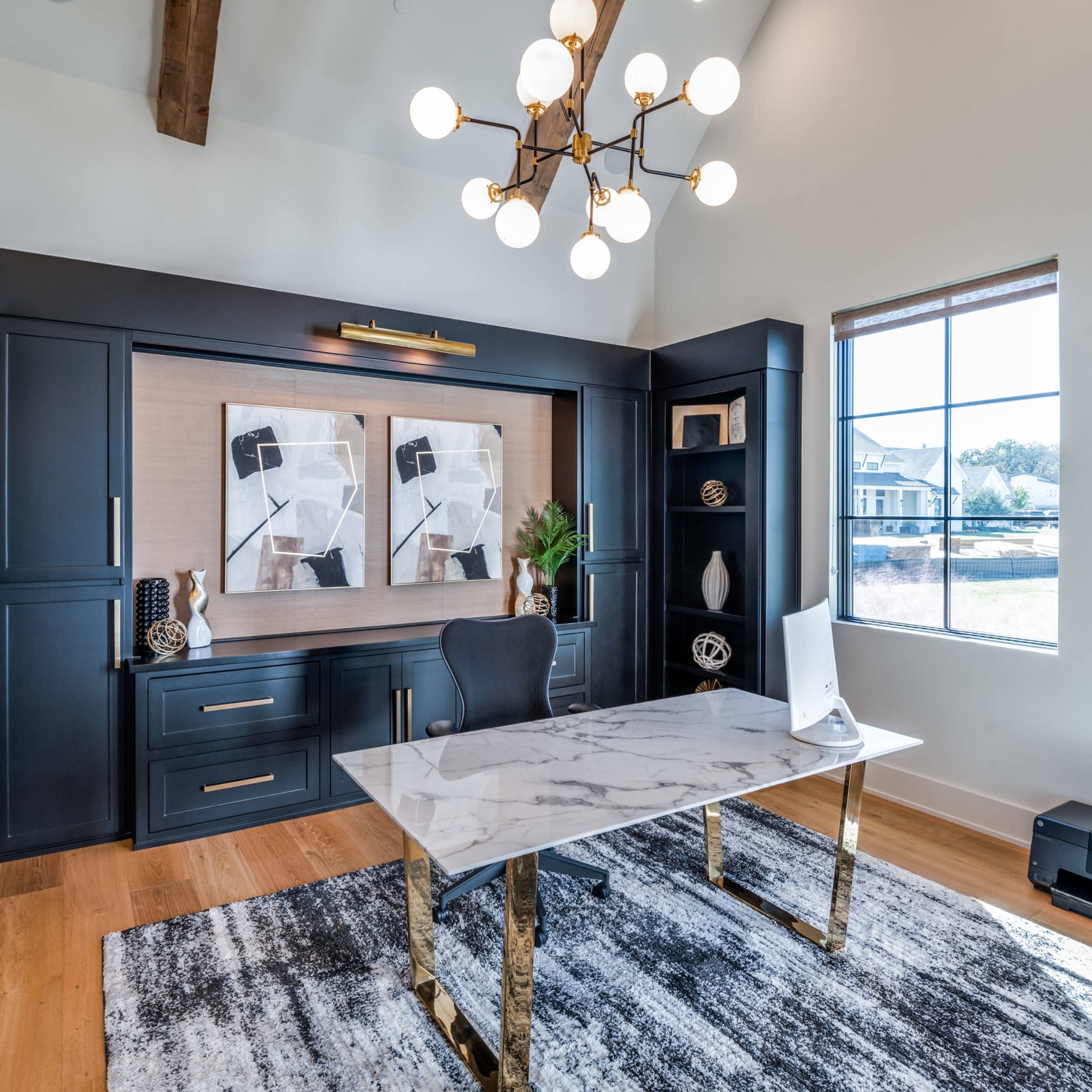 A modern office features a marble-topped desk, a black chair, a stylish chandelier, and artwork displayed on dark cabinetry against a bright wall.