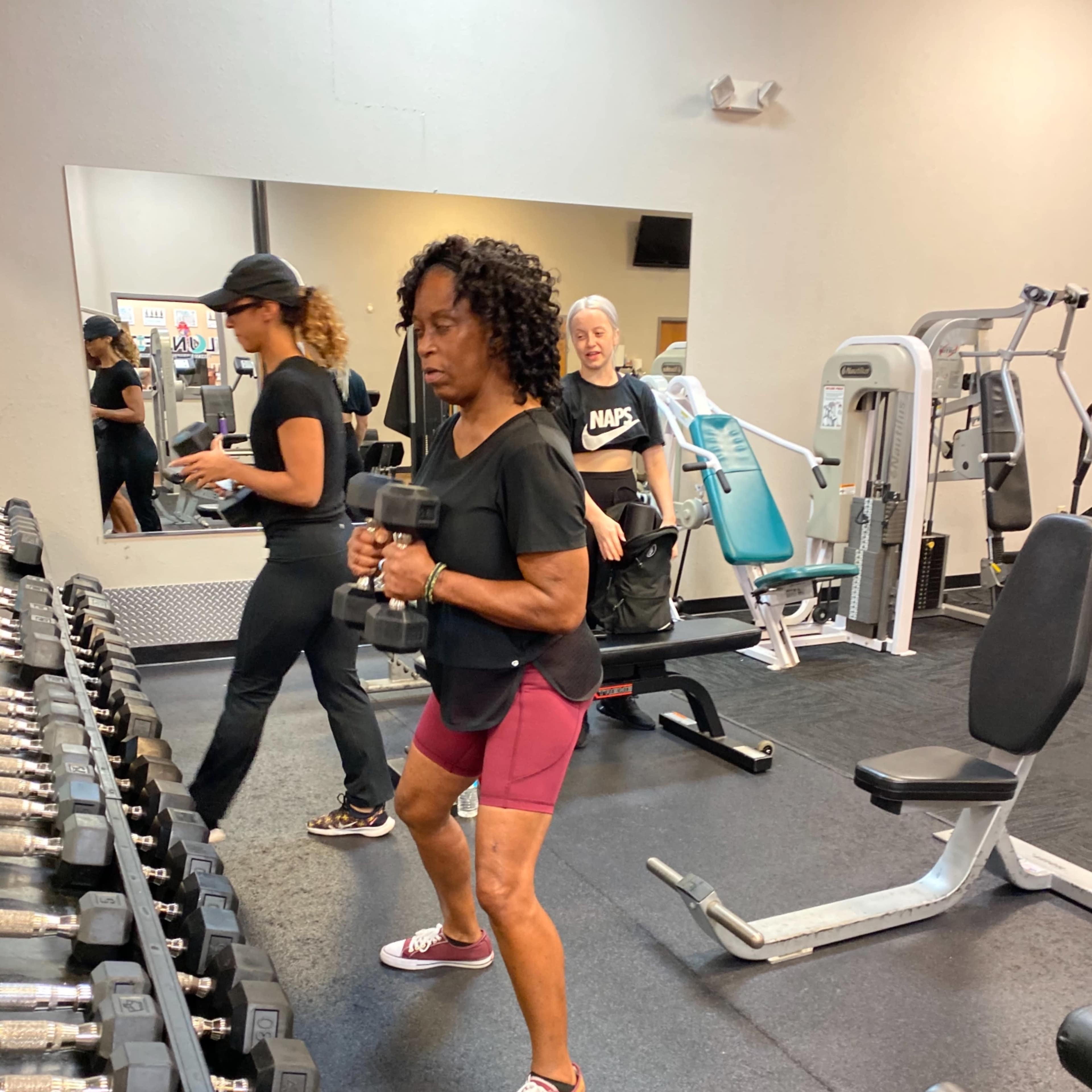 A woman in a black shirt and maroon shorts is performing a weighted exercise with dumbbells in a gym, while another woman and a third figure are engaged in different activities nearby.
