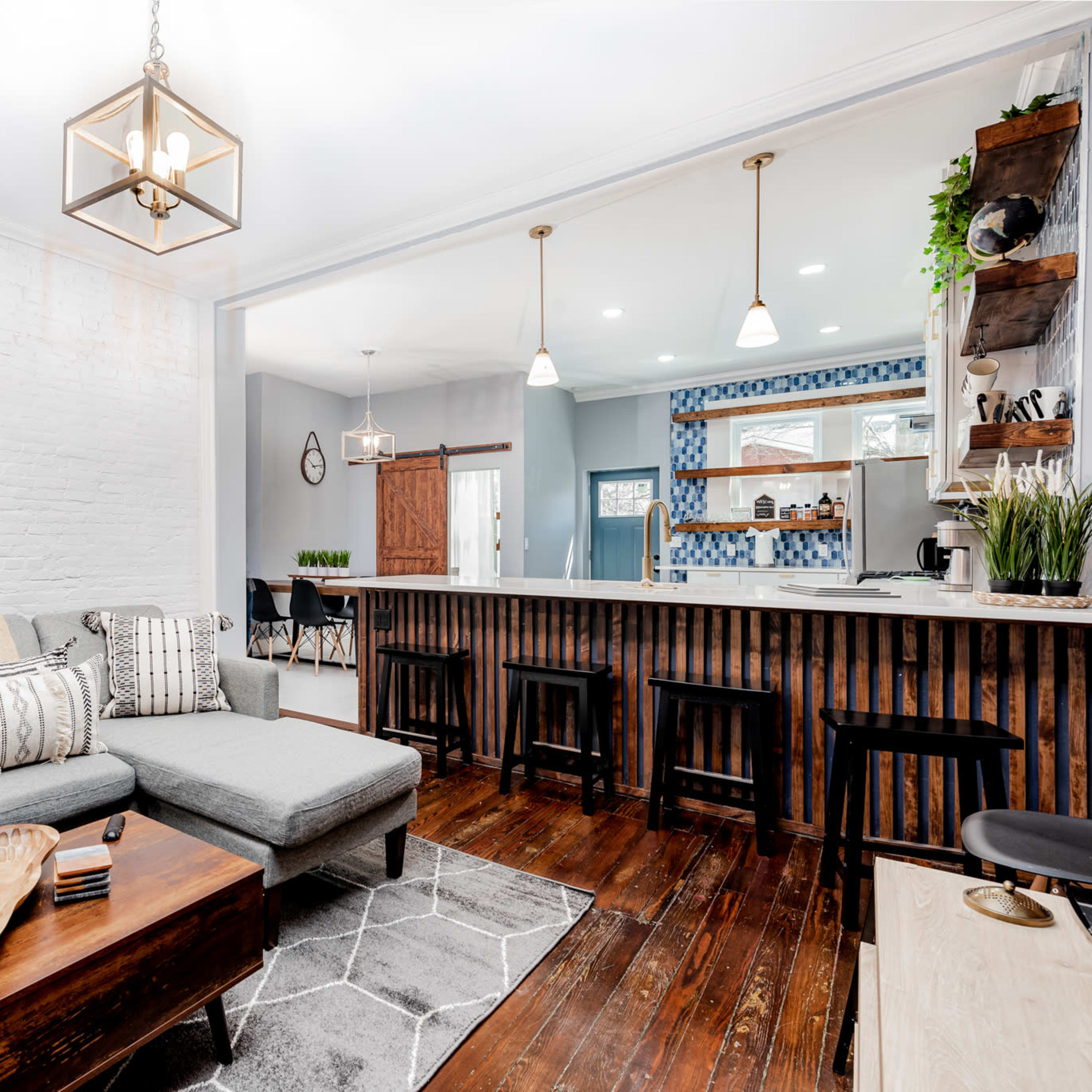 A modern living room features a gray sectional sofa, a wooden coffee table, and an open kitchen with a bar and blue tile backsplash.