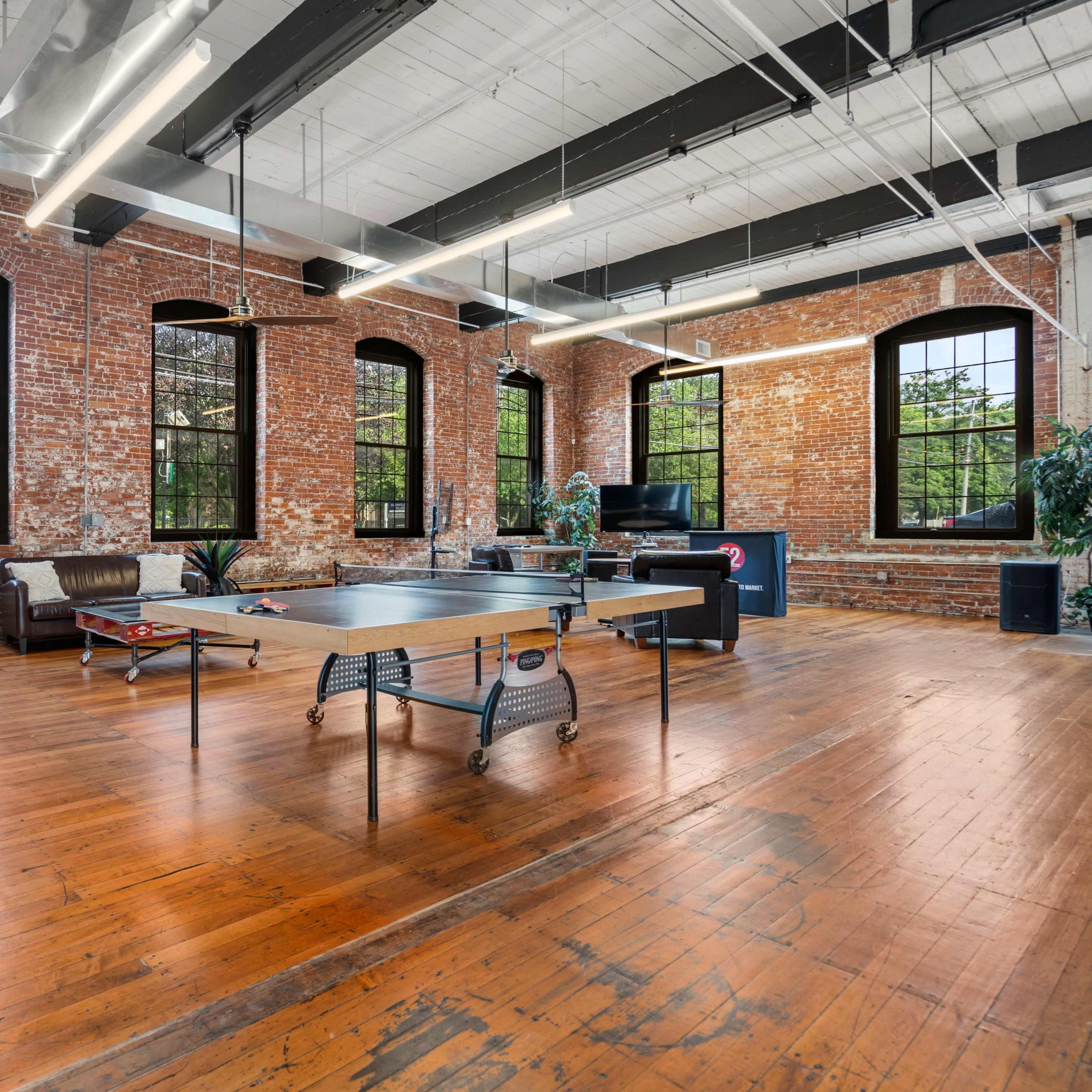 A spacious loft with exposed brick walls and large windows features a ping pong table in the center and hardwood flooring.