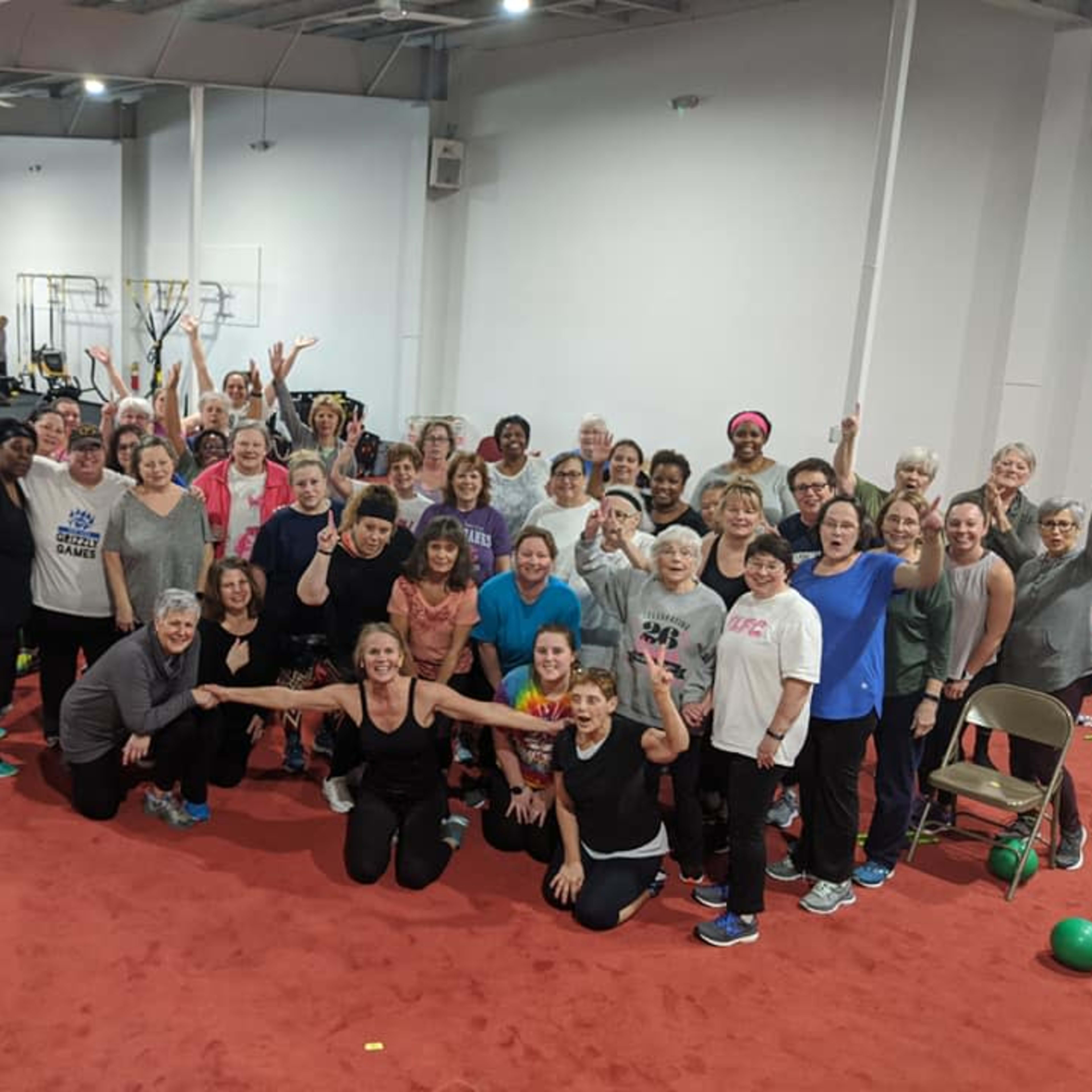 A large group of women of various ages poses together in a fitness center, with exercise equipment visible in the background.