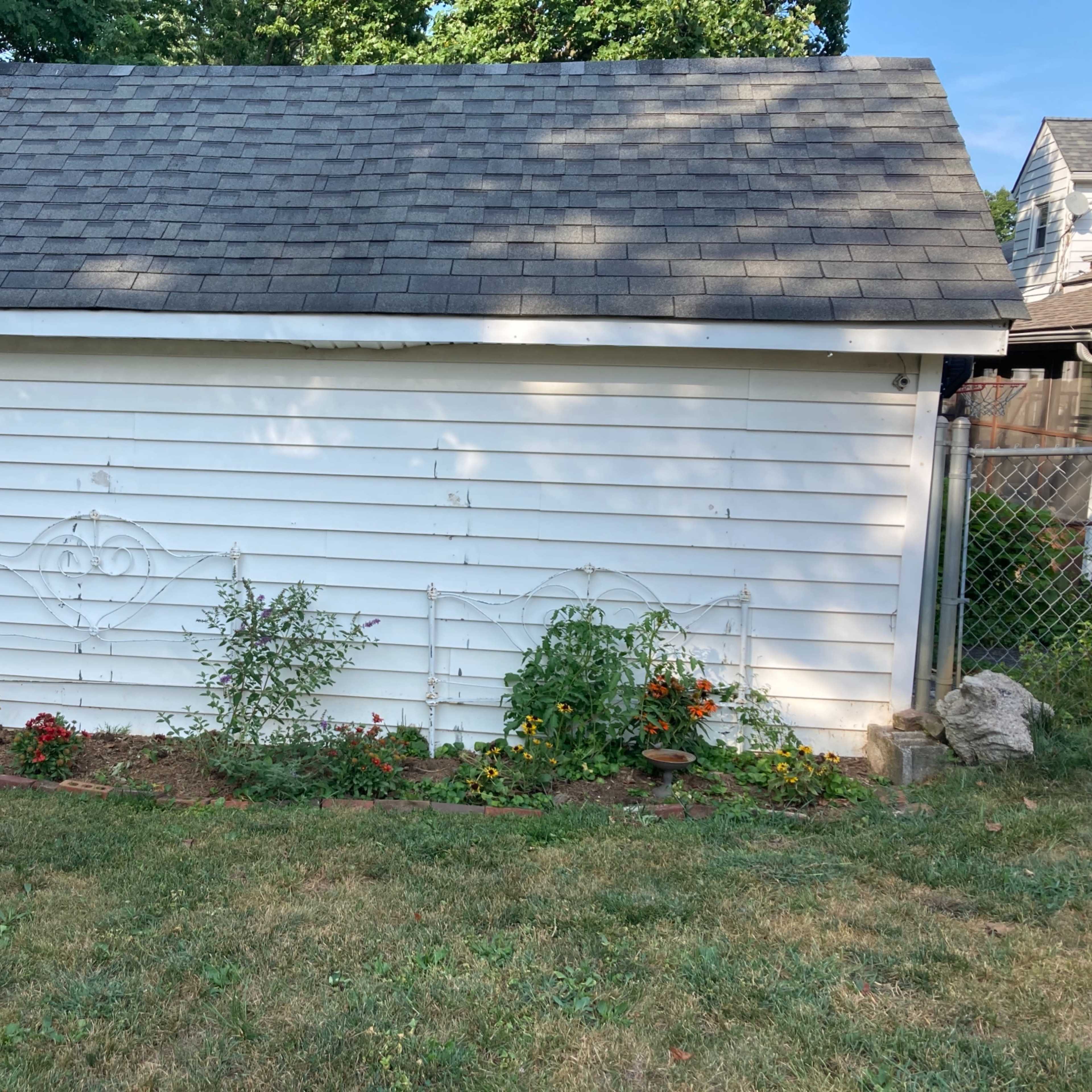 A white shed with a gray roof has a flowerbed lined with various plants and flowers in front of it.