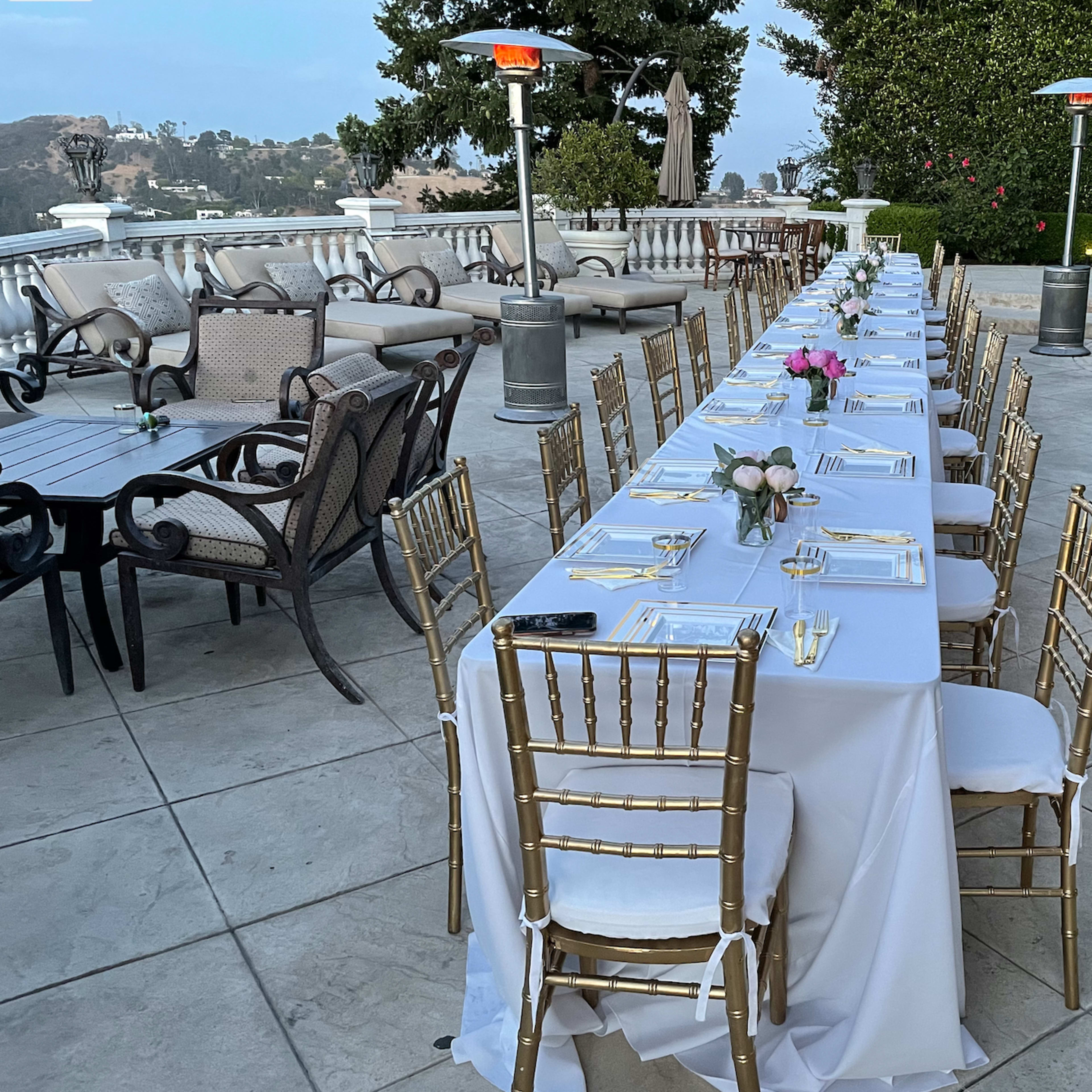 A long, decorated dining table is set outdoors with gold chairs and a view of surrounding greenery and a pool.