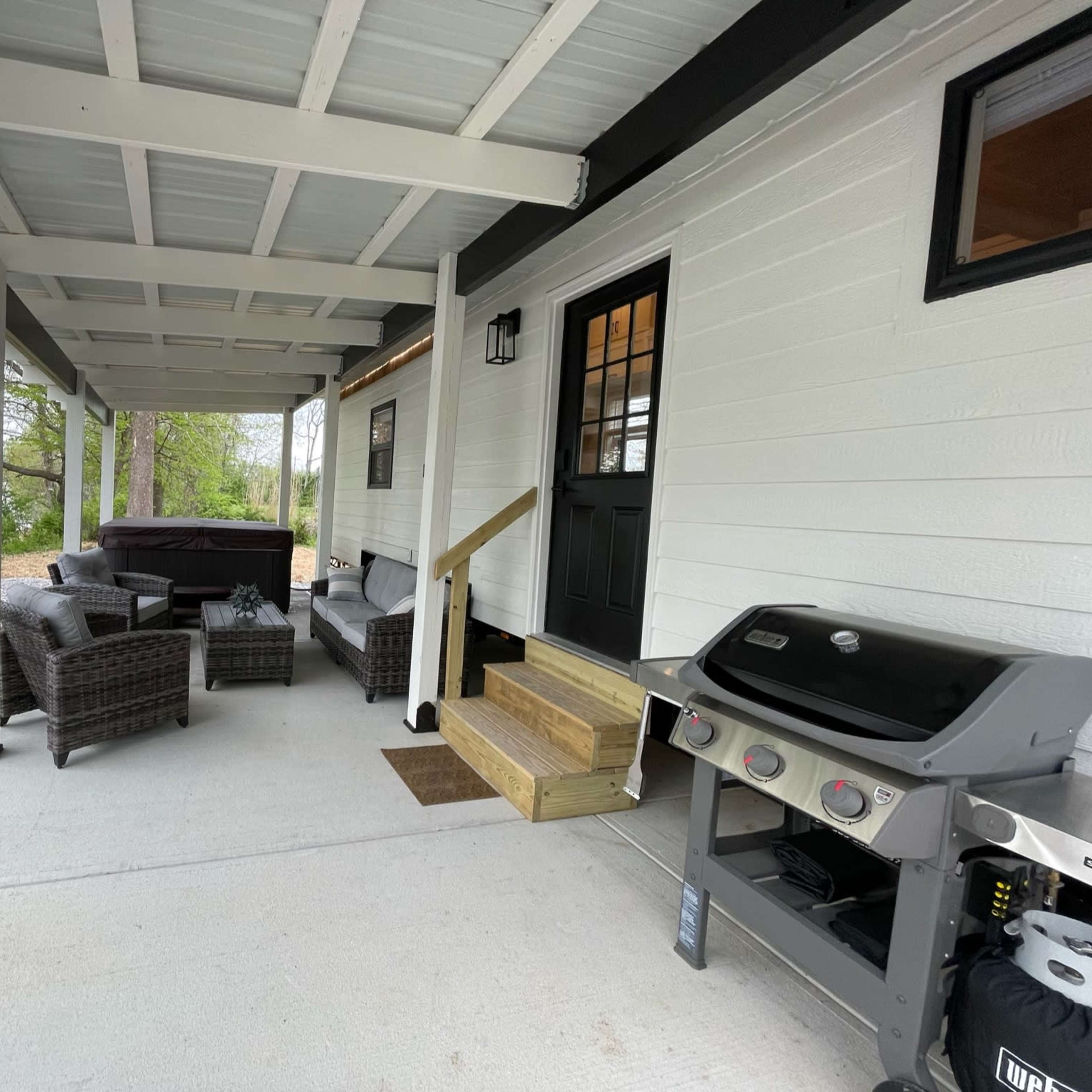 A covered outdoor patio with seating, a barbecue grill, and steps leading to the entrance of a house.