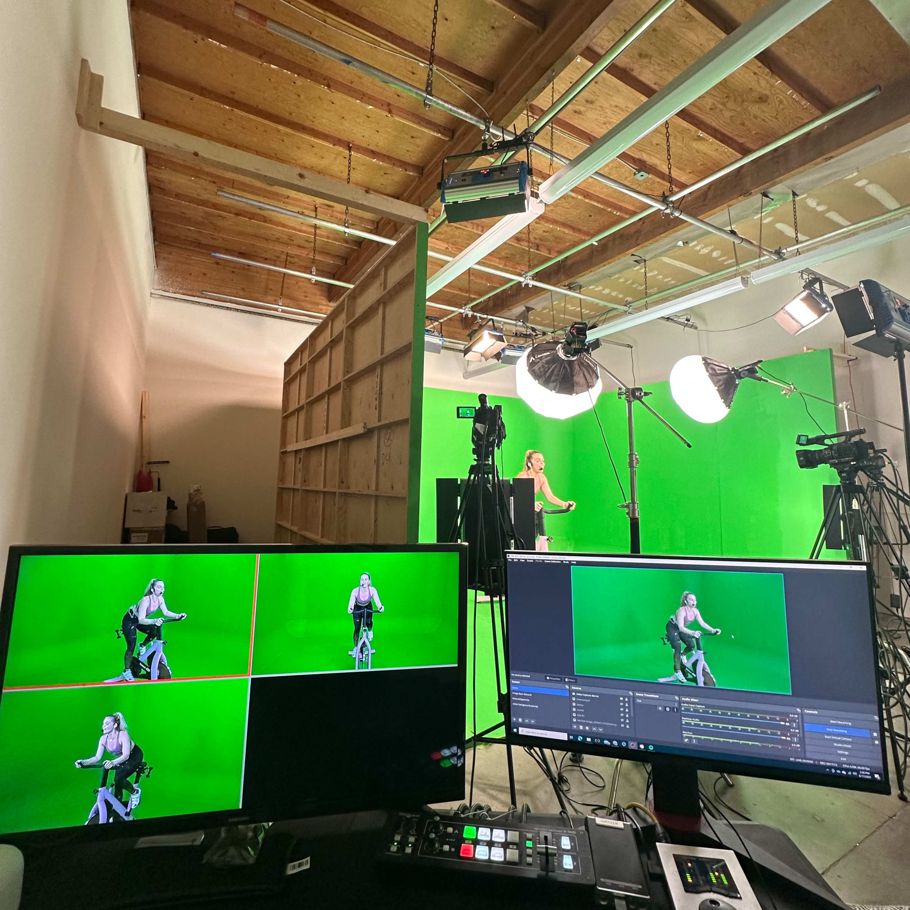 A green screen studio setup with multiple cameras and monitors displaying a person on a stationary bike in front of a green backdrop.
