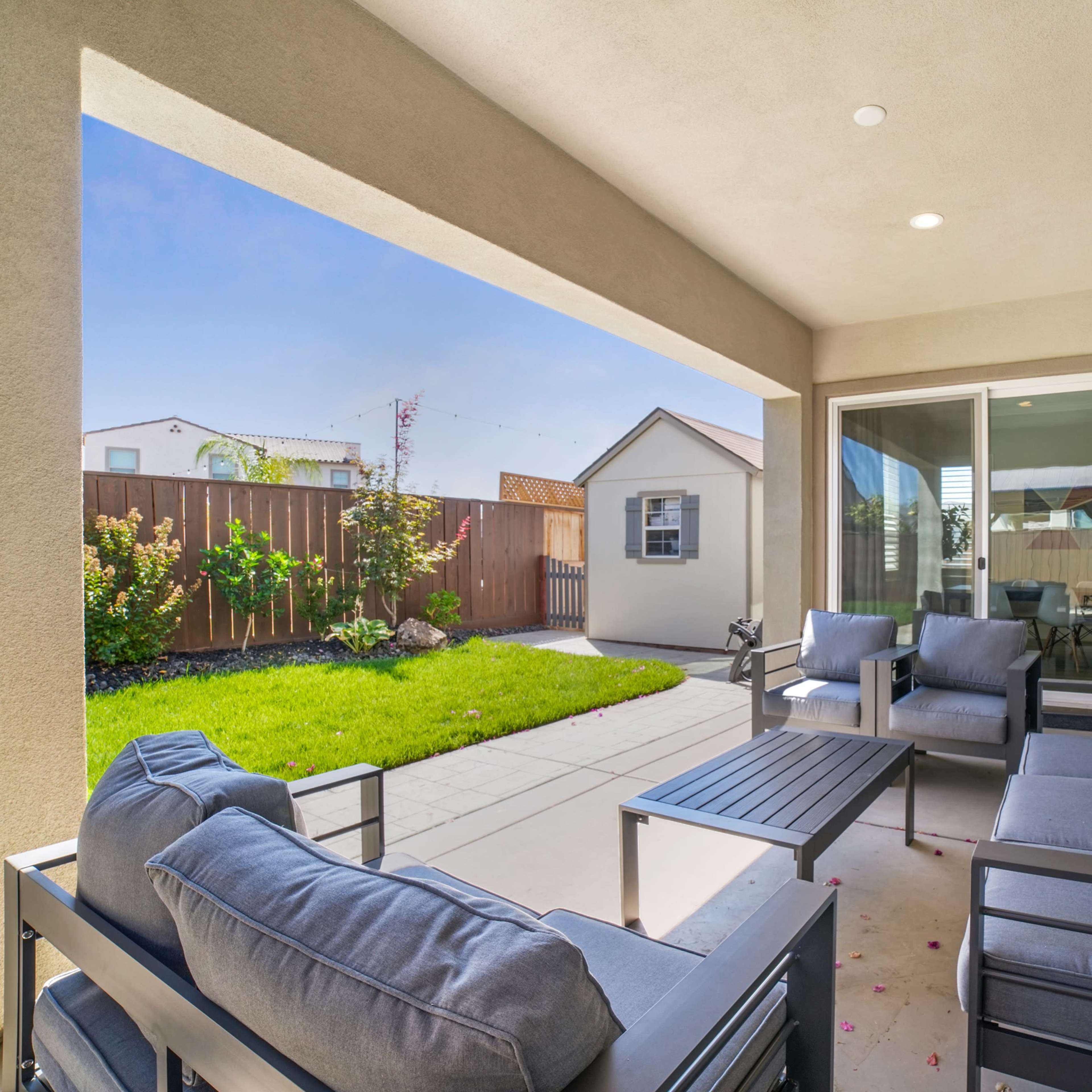 A covered patio features a seating area with cushioned chairs and a coffee table, overlooking a neatly landscaped backyard with a shed.