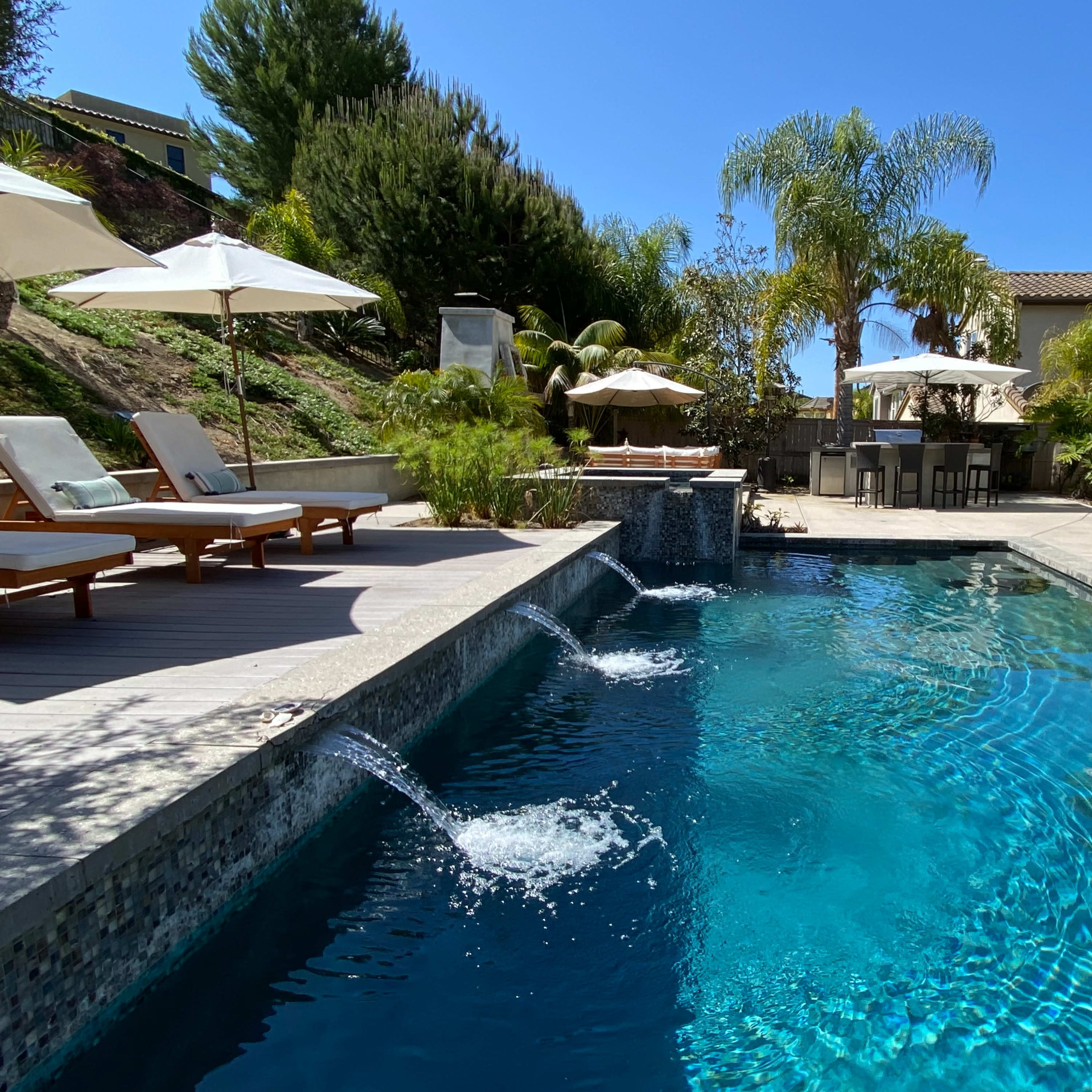 A residential backyard with a swimming pool featuring waterfall fountains, surrounded by lounge chairs, parasols, and a dining area.