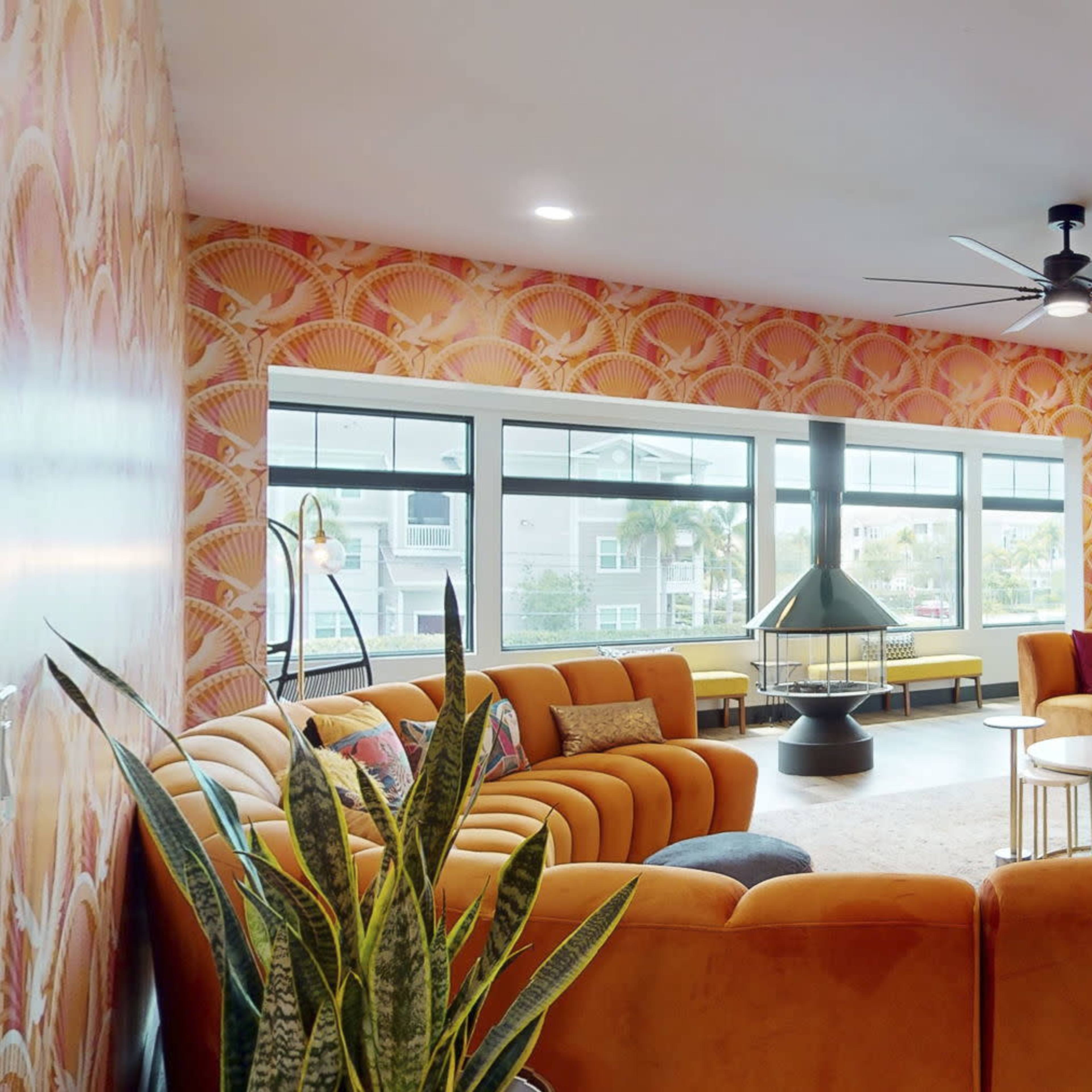 A brightly colored lounge features a circular arrangement of orange sofas around a central coffee table, with large windows letting in natural light.