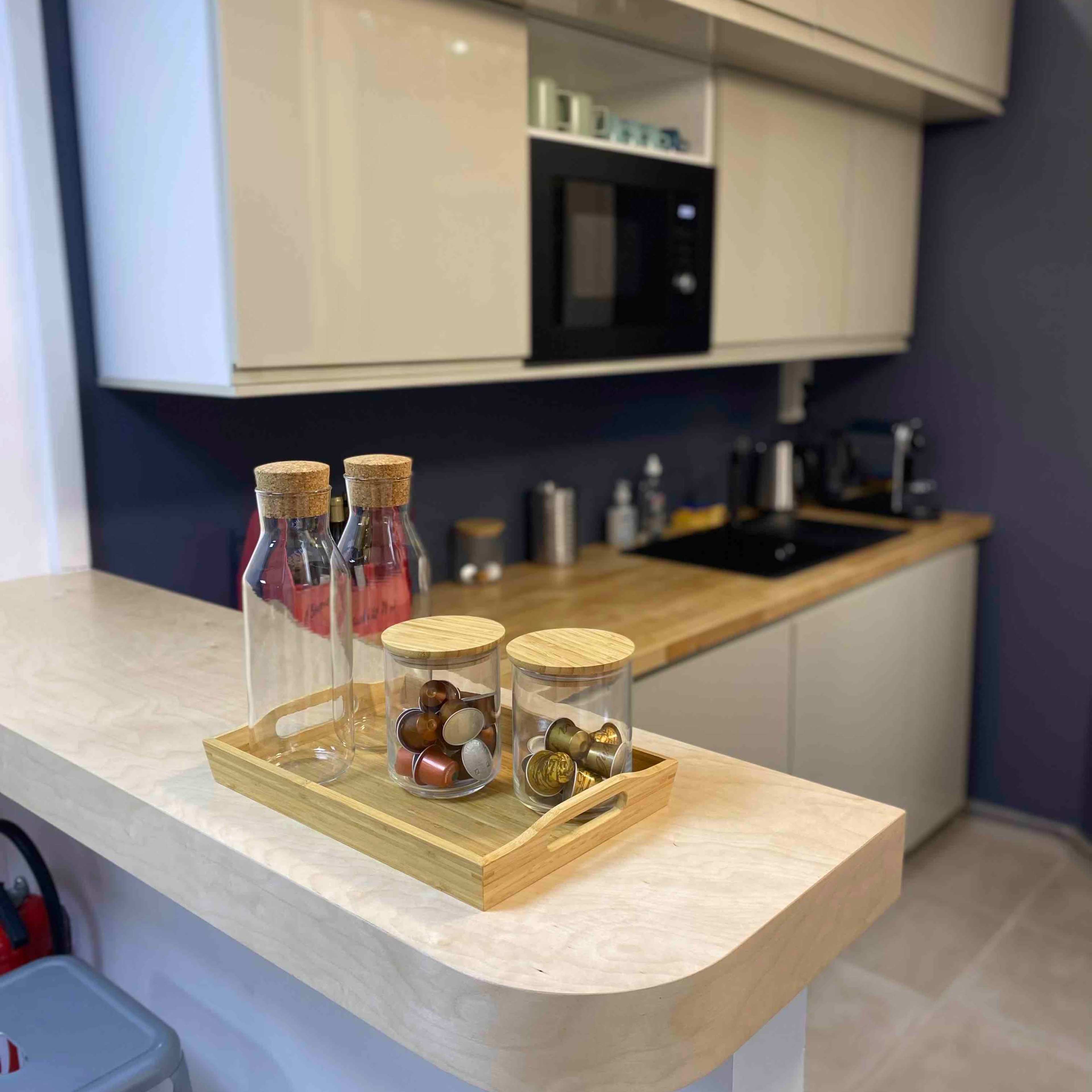 A modern kitchen with a light-colored countertop, a wooden surface, and three glass containers with wooden lids on a tray, while a microwave and other appliances are visible in the background.