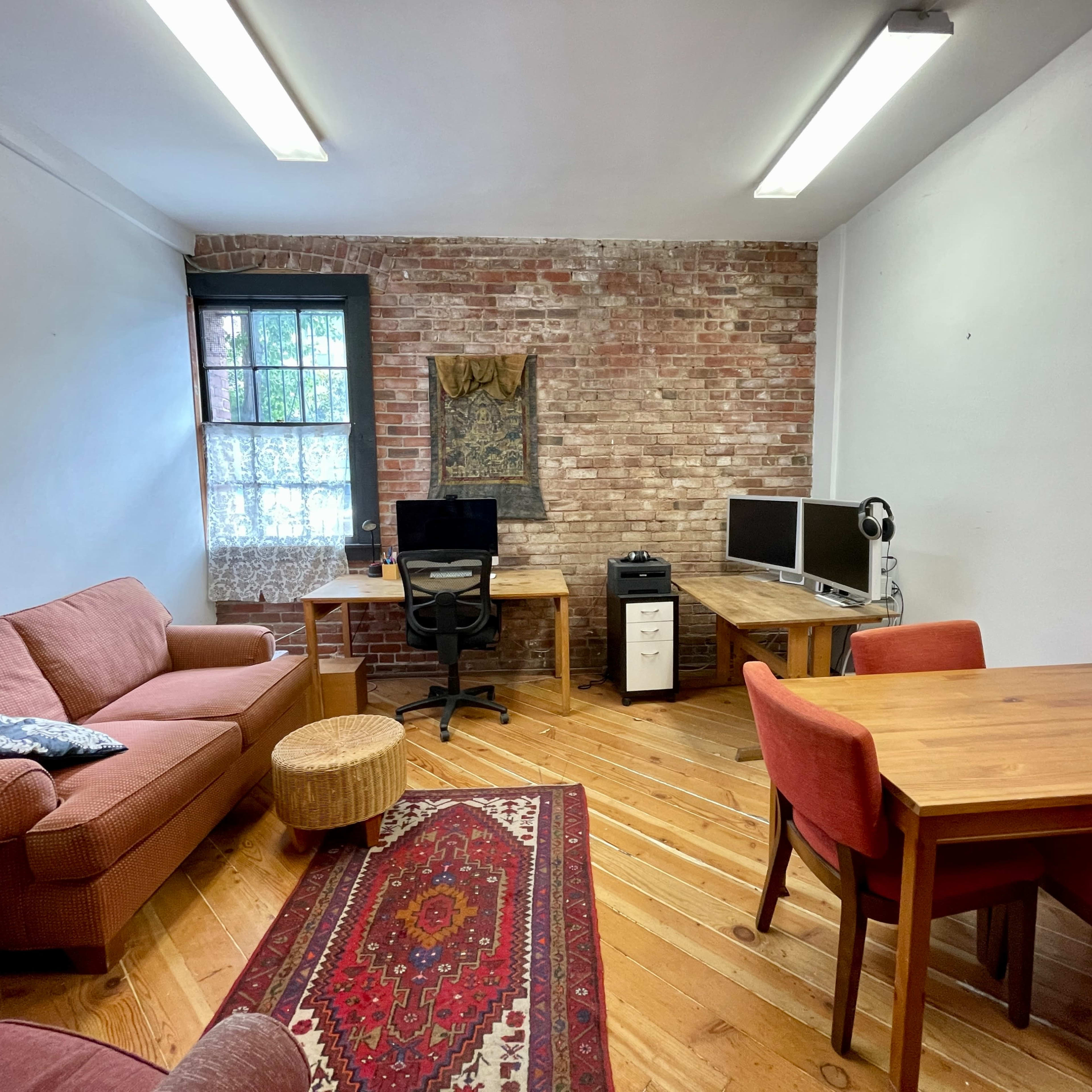 A room with exposed brick walls, wooden flooring, a sofa, a desk with a computer chair, two computer monitors, a filing cabinet, a dining table with red chairs, and a decorative rug.
