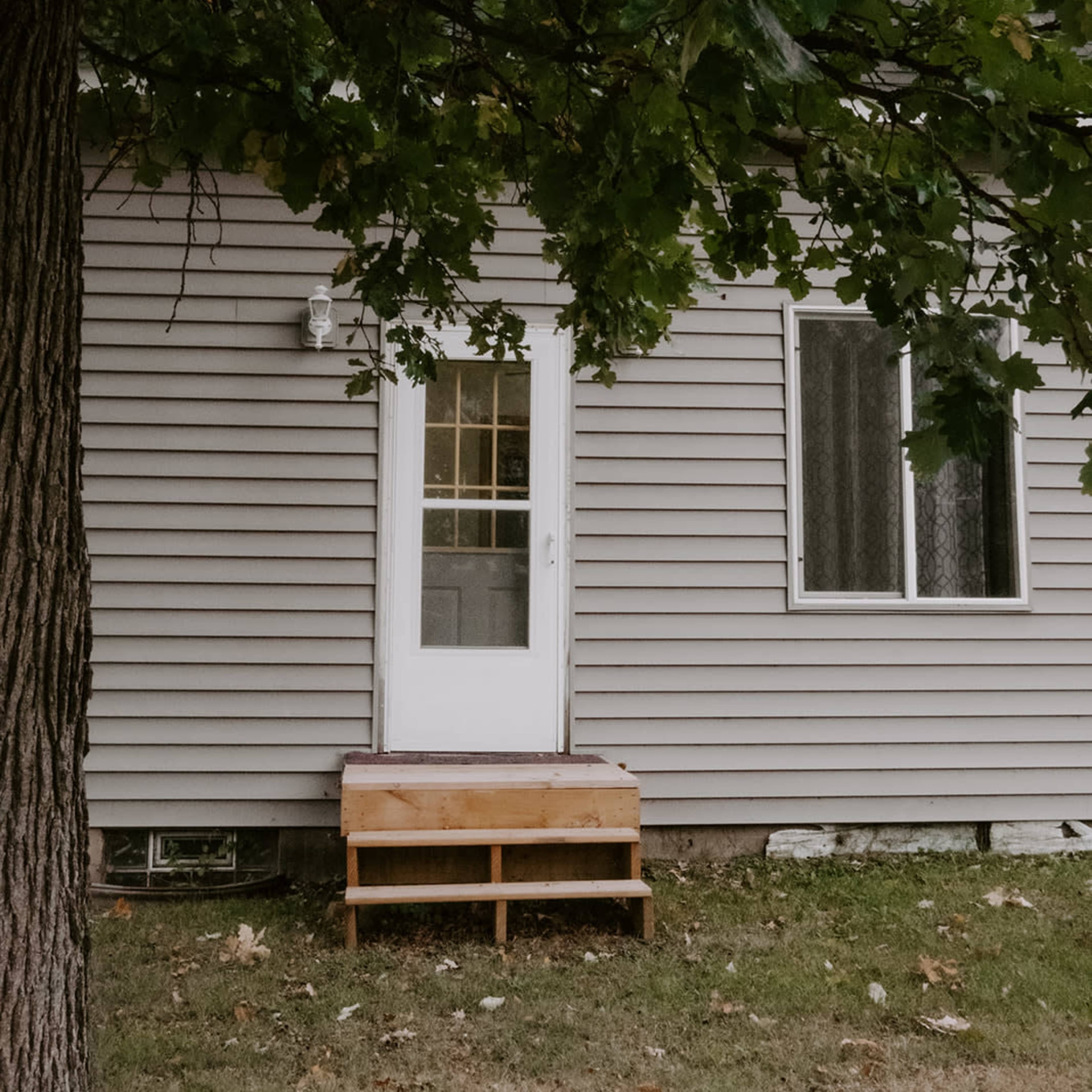 A gray house with a white door and wooden steps, partially obscured by a tree in the foreground.